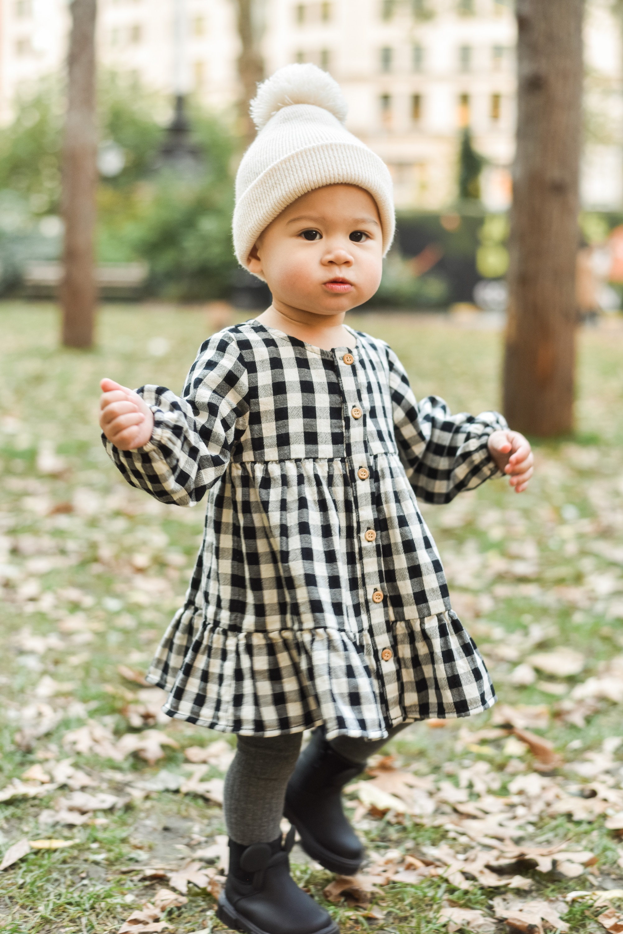 Toddler in white knit beanie and black-and-white gingham dress stands in a park surrounded by fallen leaves and bare trees, with blurred urban buildings behind