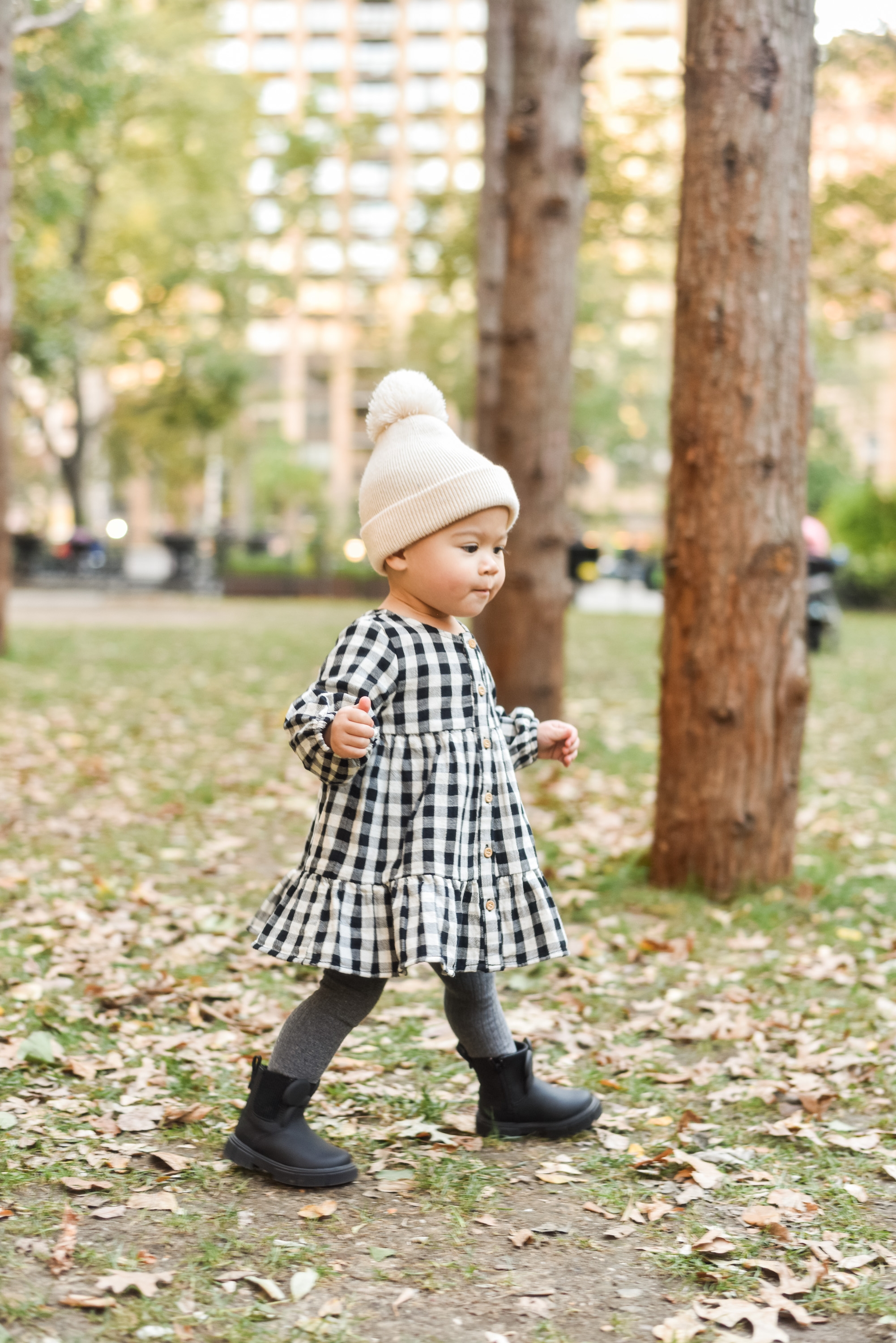 A toddler in a black-and-white gingham dress and cream knit beanie walks across fallen leaves in an urban park with tall trees and city buildings visible in the soft background