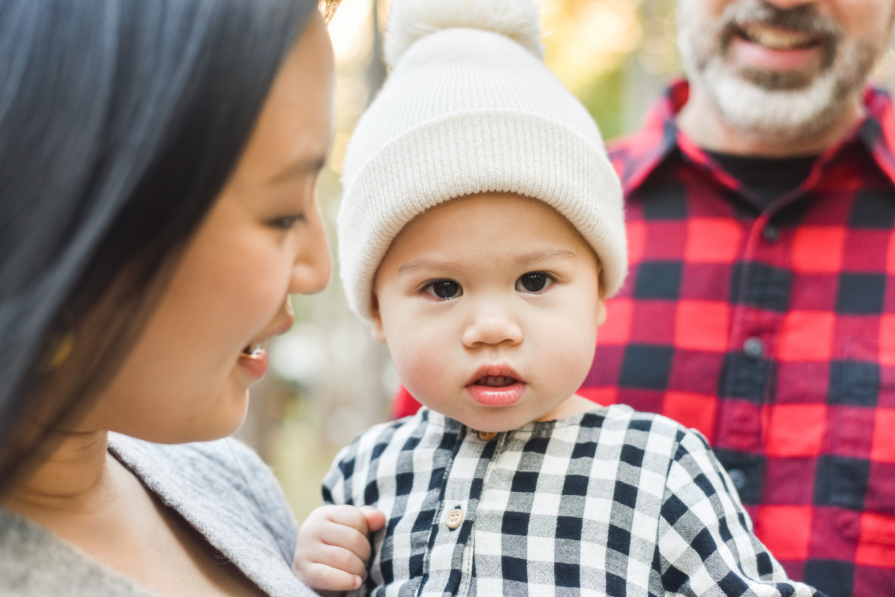 A toddler in a white beanie and checkered shirt gazes directly at the camera while held by a woman, with a man in a red plaid shirt visible in the blurred background