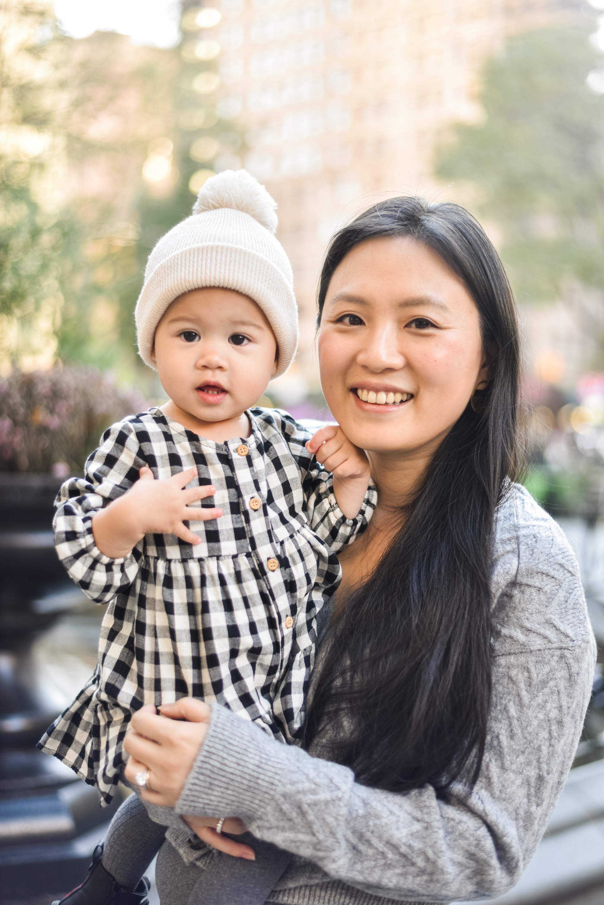 A smiling woman holds a toddler wearing a white knit hat and checkered shirt in an urban park setting with blurred autumn foliage background