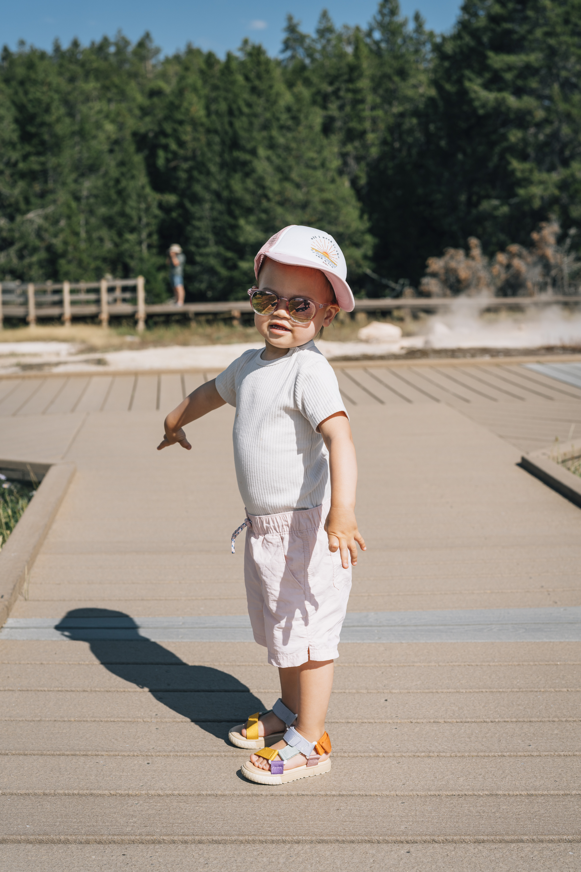 A young child wearing a white cap and sunglasses stands on a concrete boardwalk, with evergreen forest and geothermal features visible in the bright daylight background