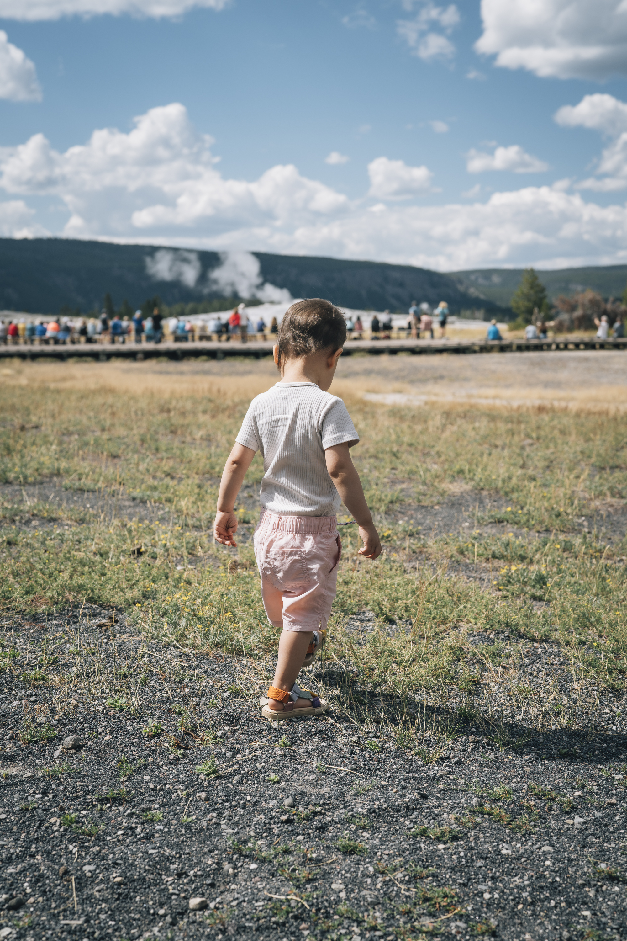 A young child in a white shirt runs across a sparse gravel field toward distant geysers and forested hills under a bright, cloudy sky