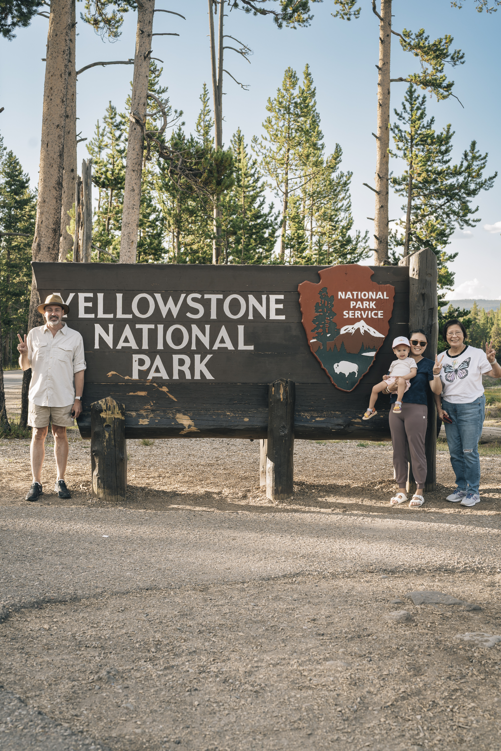 A family of four poses beside the Yellowstone National Park entrance sign on a dirt road, with tall pine trees and clear blue sky in the background