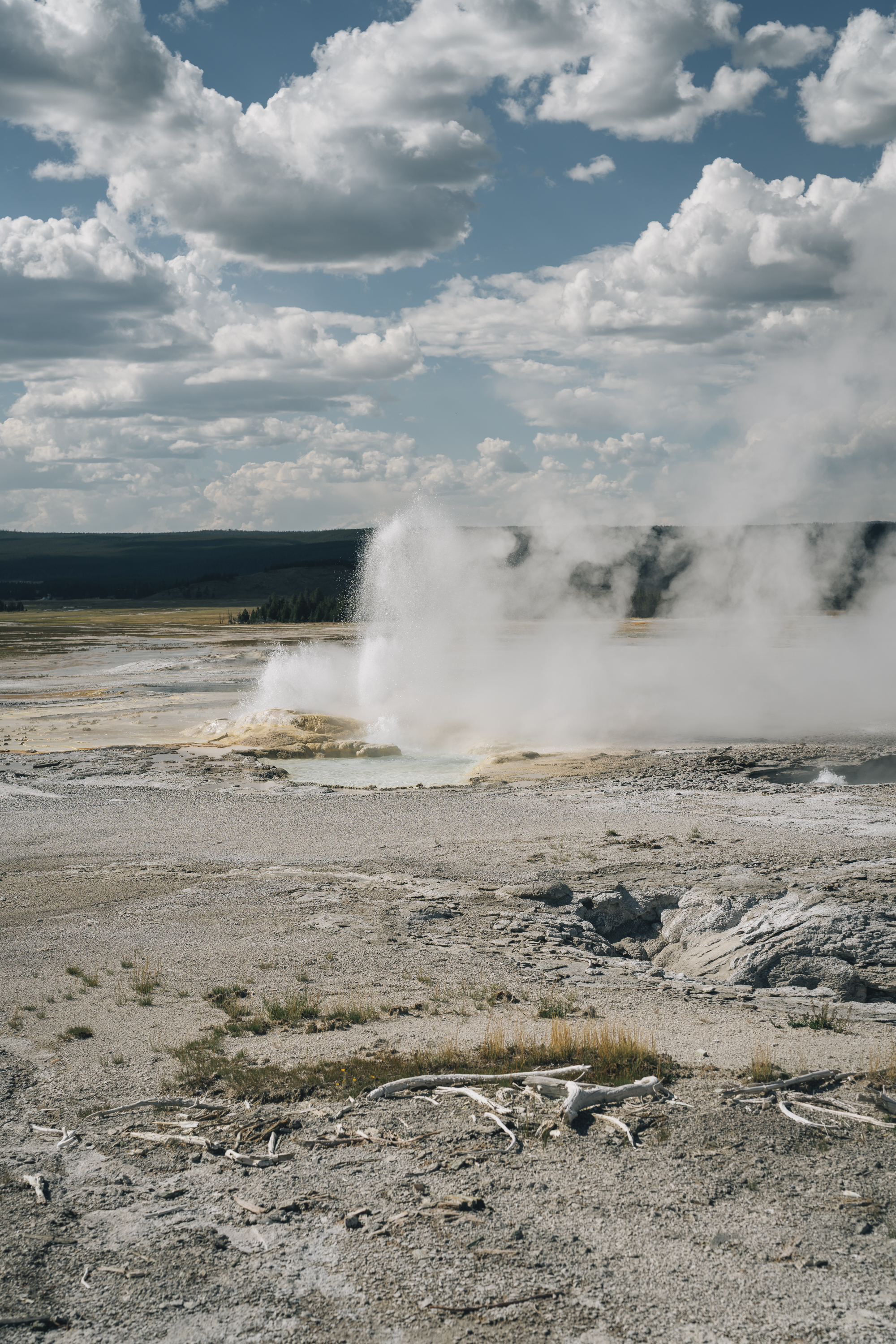 A massive geyser erupts with billowing white steam across a barren geothermal basin, with forested mountains visible in the distant background under a partly cloudy sky