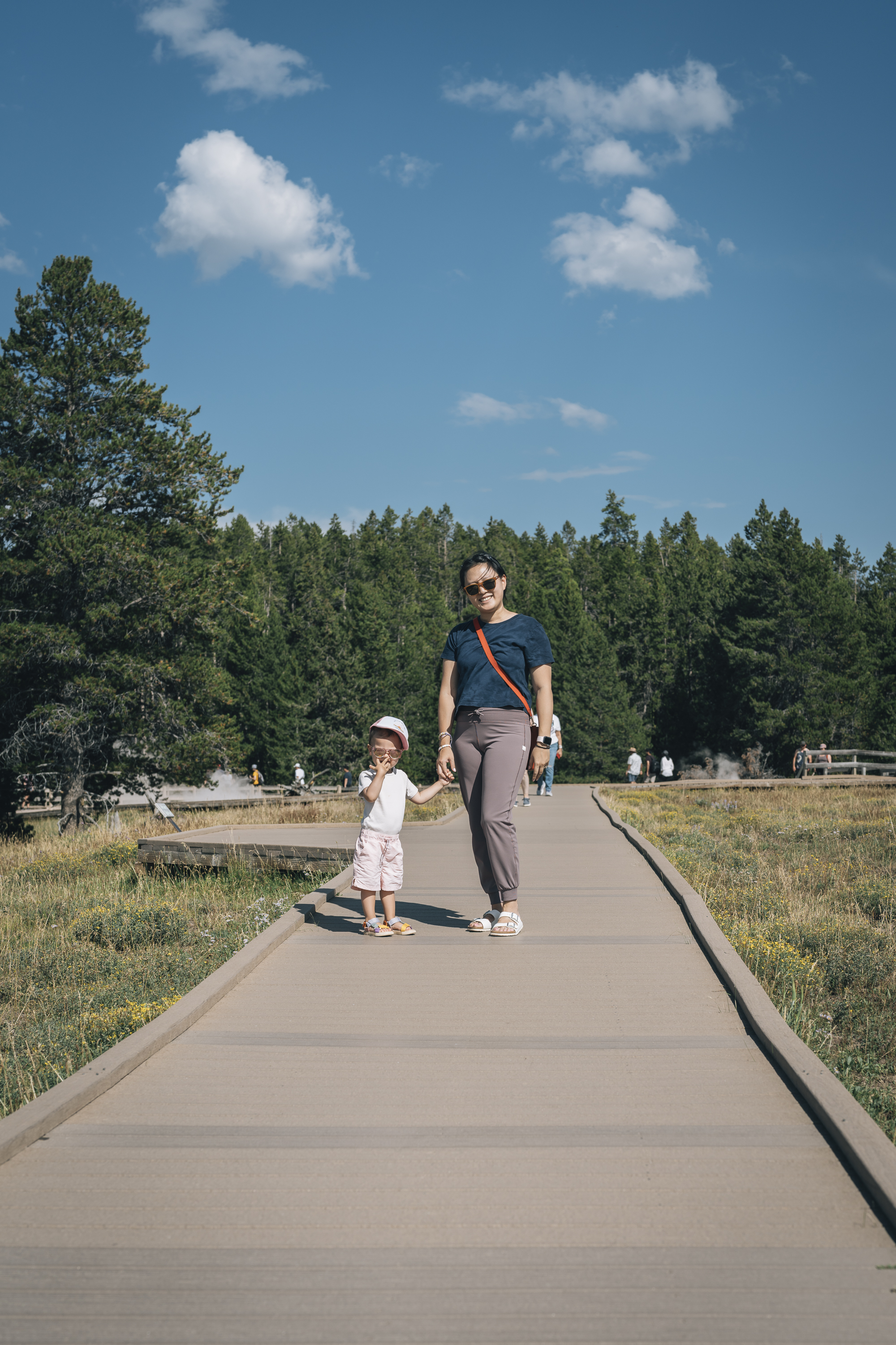 An adult and toddler walk hand-in-hand along a wooden boardwalk through a geothermal area, with a dense coniferous forest and clear blue sky in the background