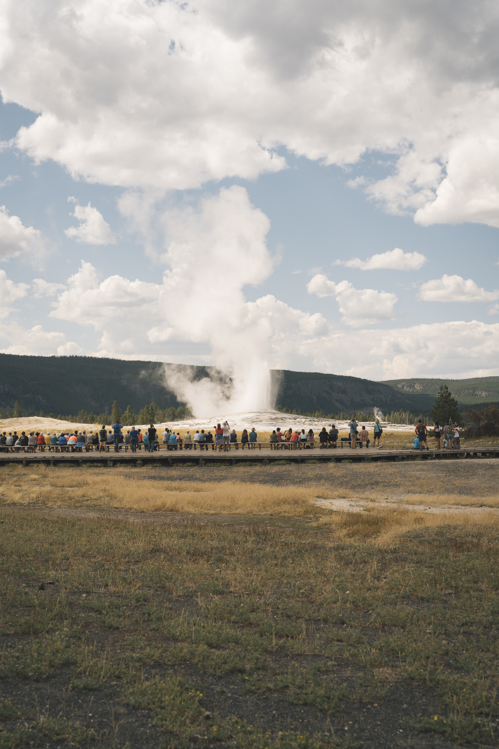 Crowds of visitors line a boardwalk to watch Old Faithful geyser erupt in a towering column of steam against a partly cloudy sky and forested hillside backdrop