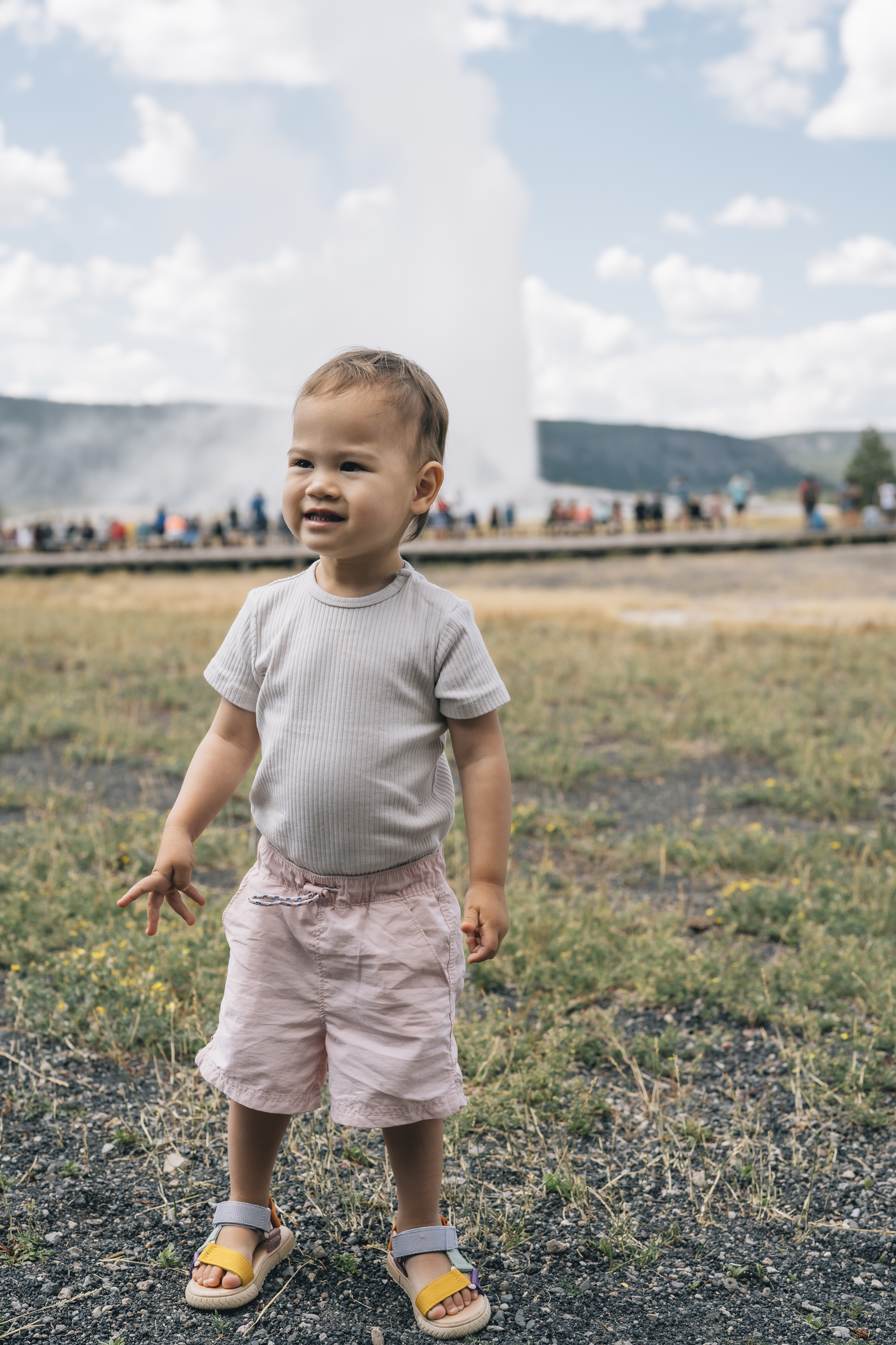 A smiling toddler in a gray shirt and pink shorts stands on a gravel path with geysers and mountains visible in the distant background under cloudy skies