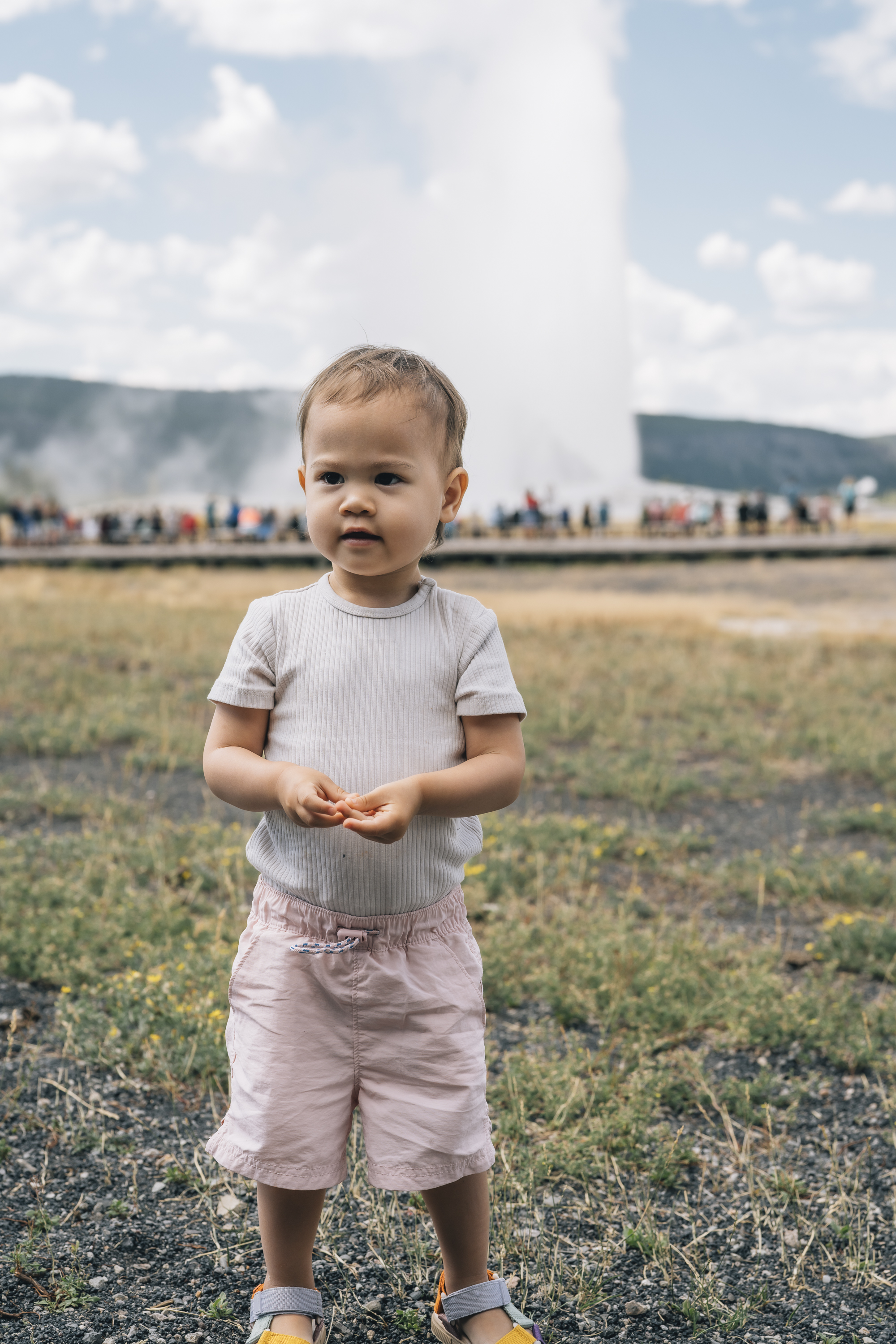 A toddler in light clothing stands in a geothermal field with Old Faithful geyser erupting in the background under a cloudy sky