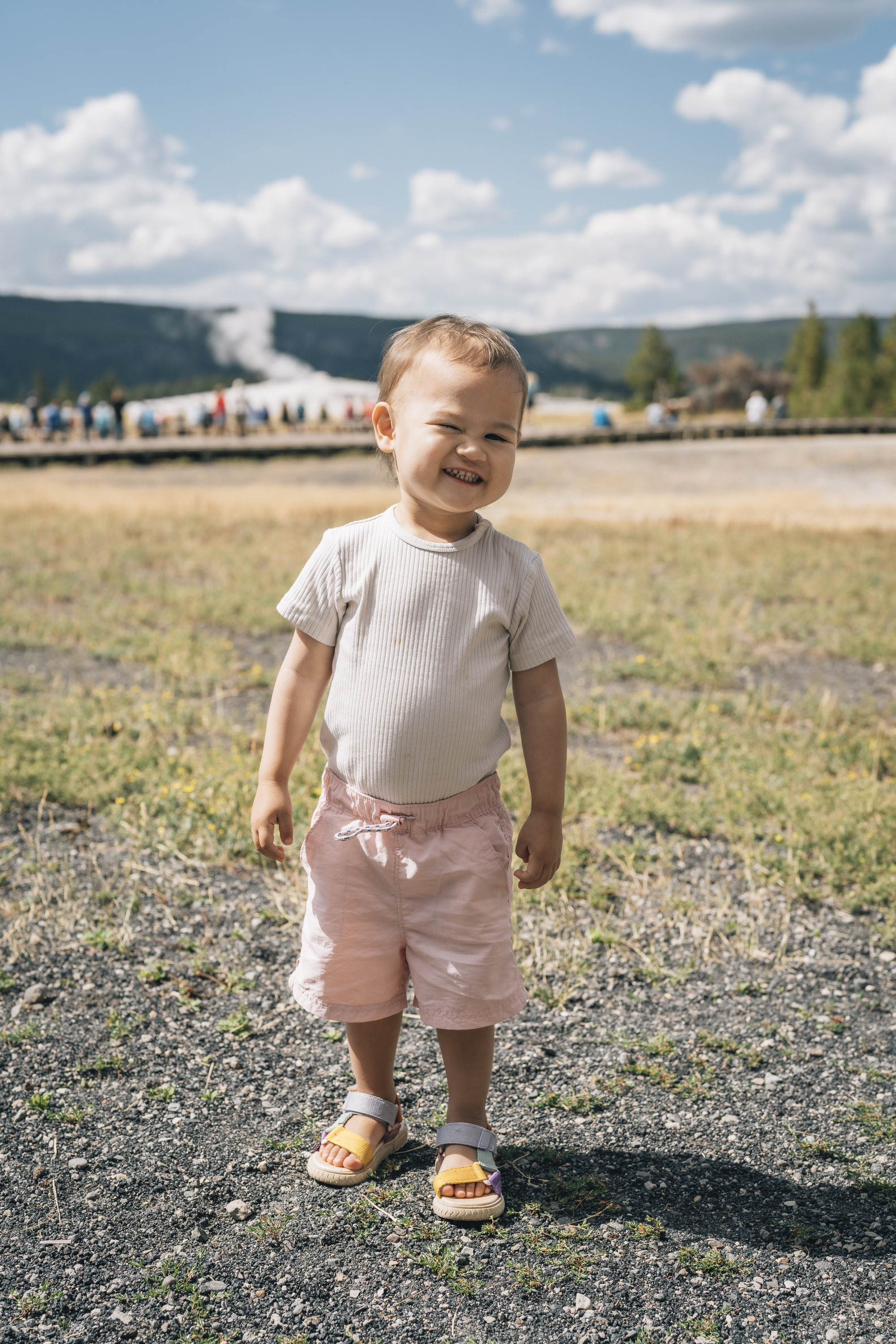 A smiling toddler in a cream shirt and pink shorts stands on a gravel path with expansive meadowland, distant mountains, and Old Faithful geysers visible under a partly cloudy sky.