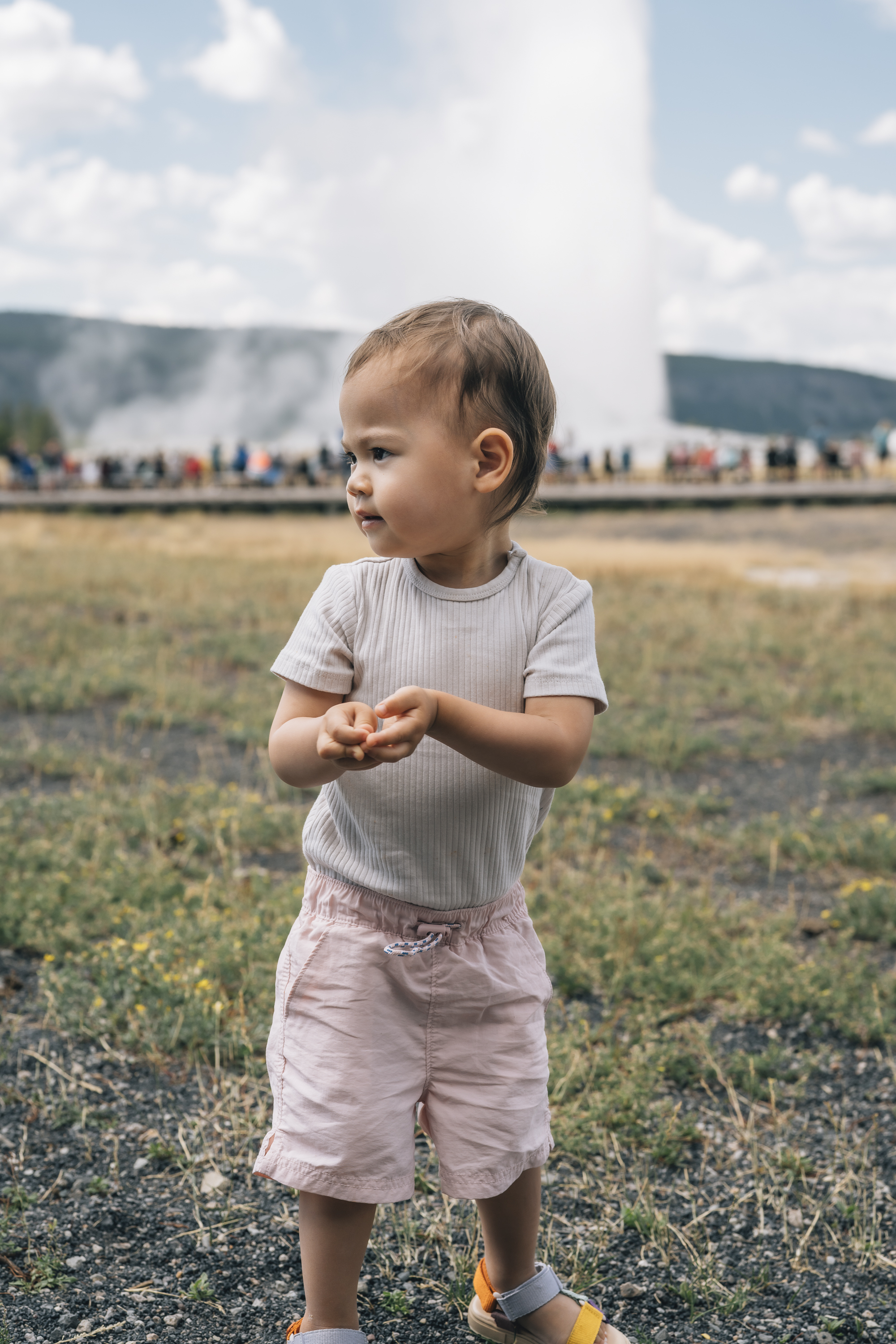 A young toddler stands alone in a grassy field facing Old Faithful geyser, with tourists and steam visible in the distant background under a partly cloudy sky