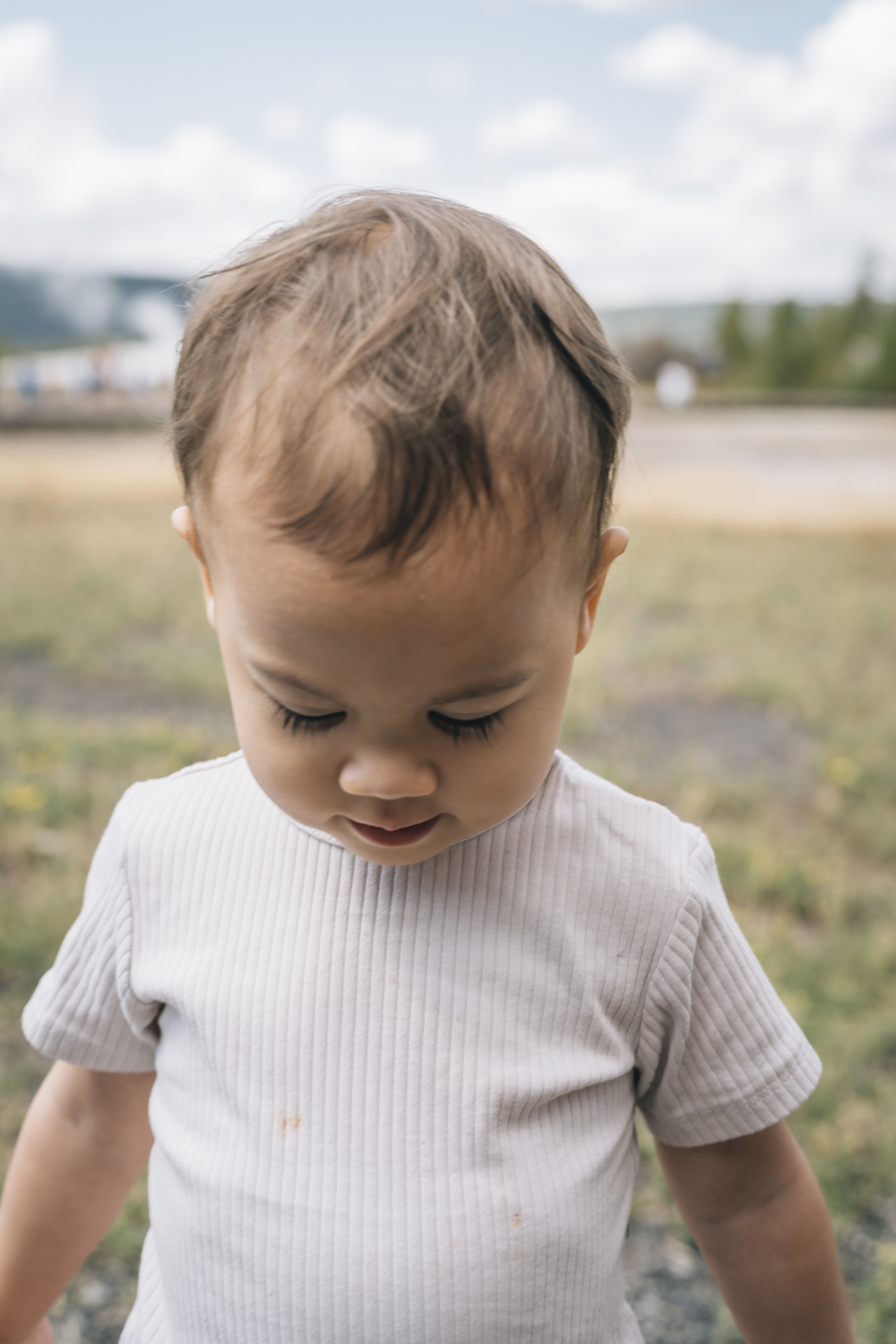A toddler in a white ribbed shirt stands in an open field with eyes downcast, mountains and evergreen trees visible in the soft, overcast daylight behind