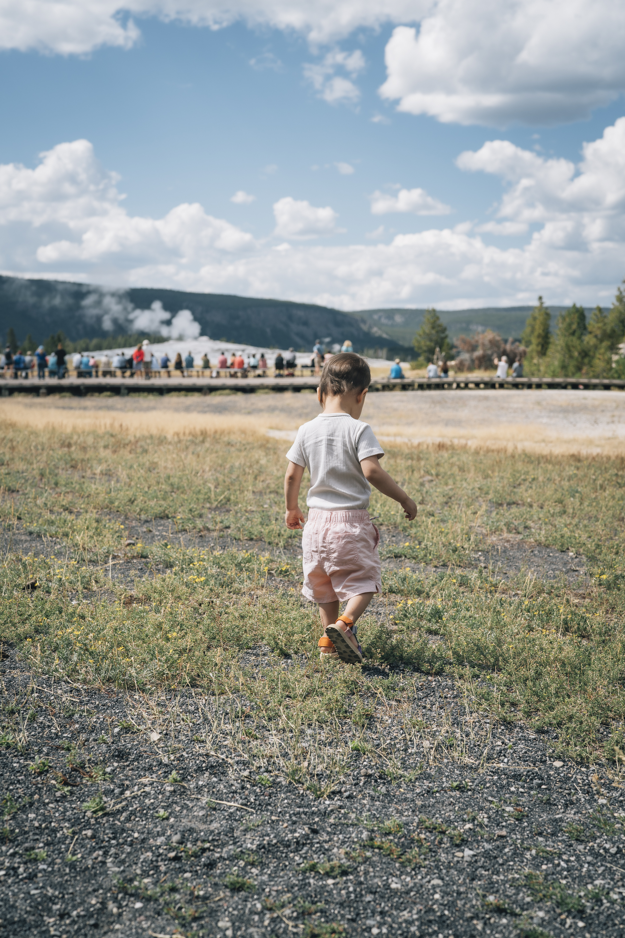 A toddler in a white shirt runs across sparse grassland toward a distant train with steam rising, surrounded by spectators and mountains under a partly cloudy sky