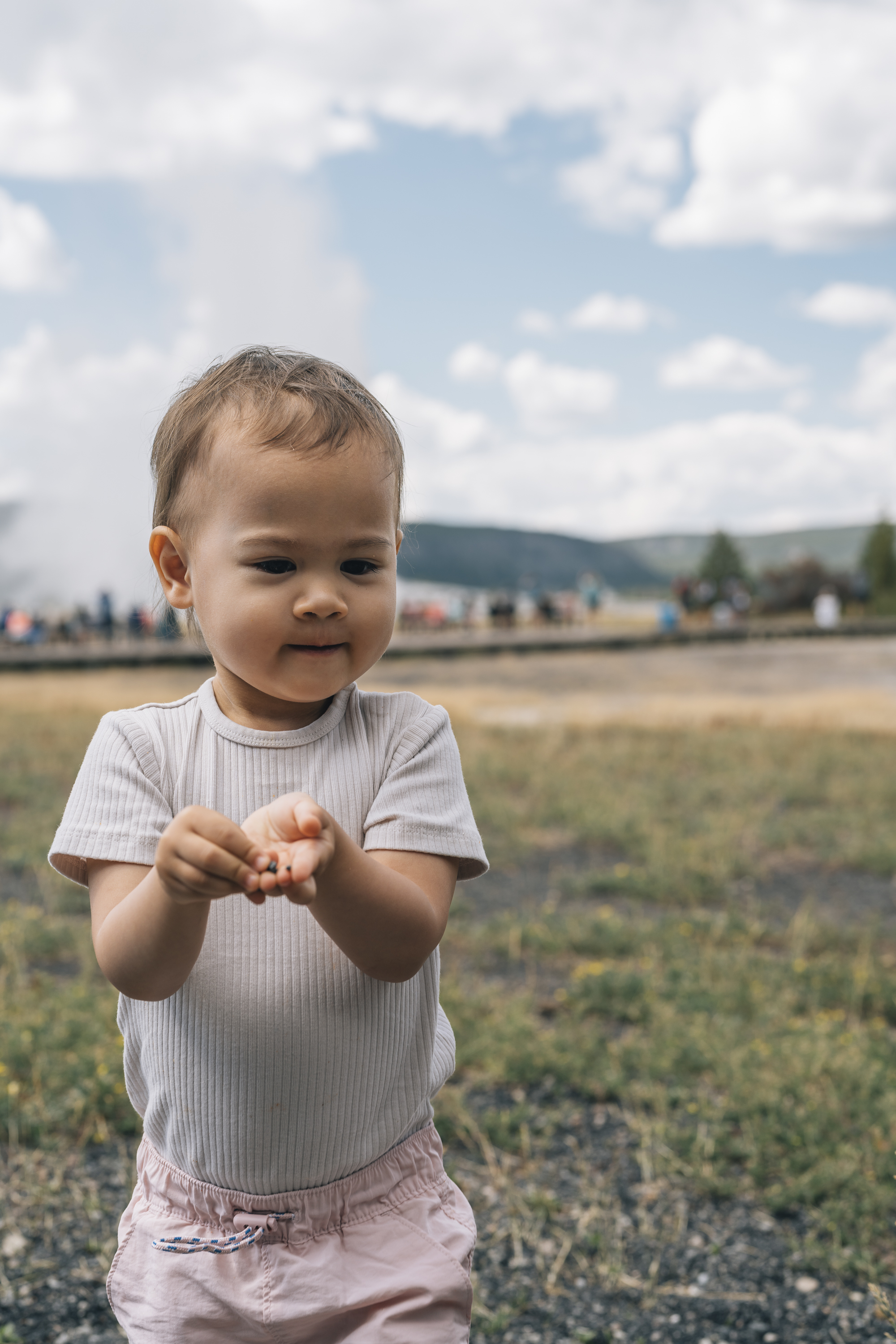 A toddler in a white ribbed shirt stands in an open field with mountains and scattered trees visible under a partly cloudy sky