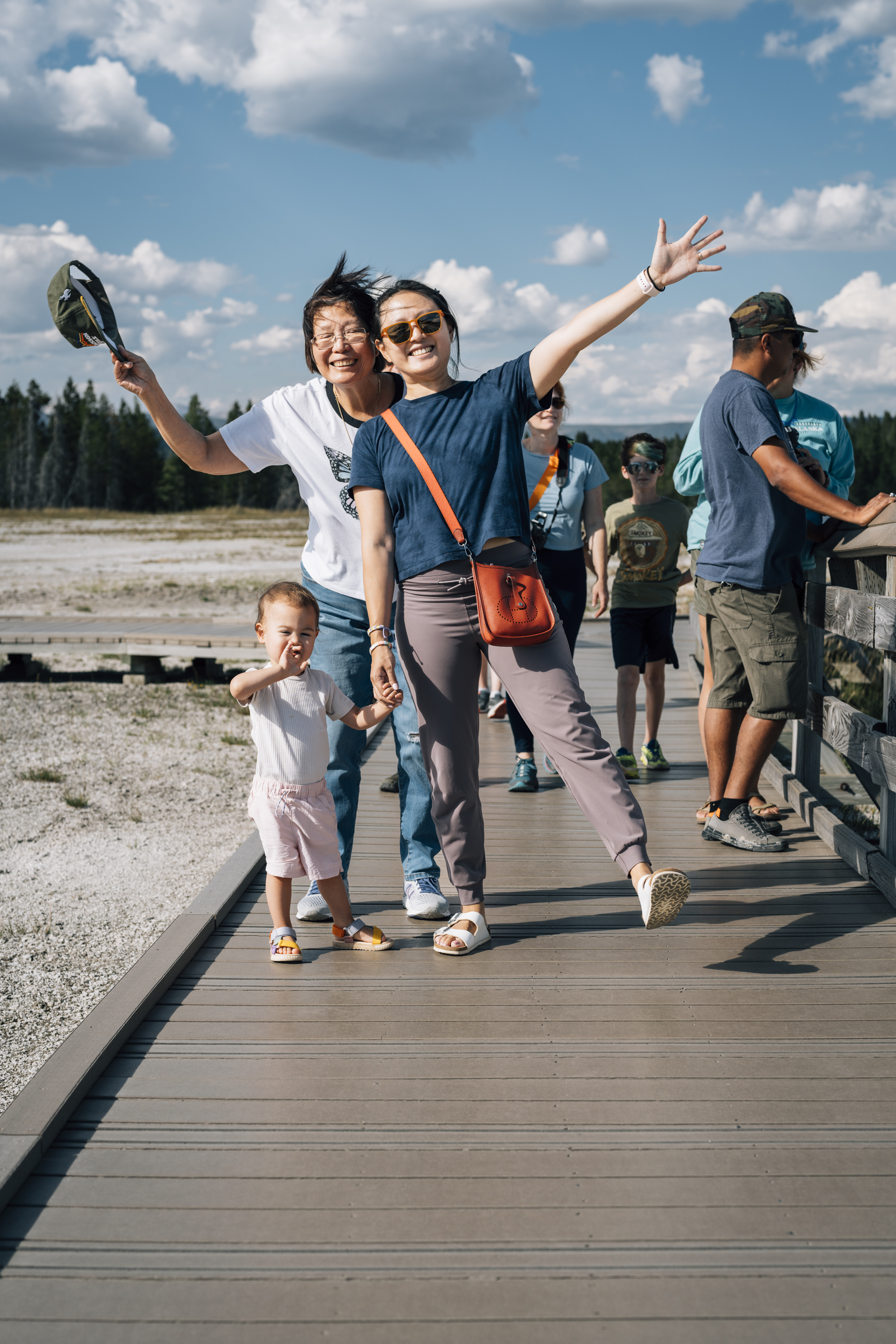 A woman with raised arm celebrates on a wooden boardwalk with a young child beside her, while other visitors stroll nearby under blue skies and distant evergreen forests