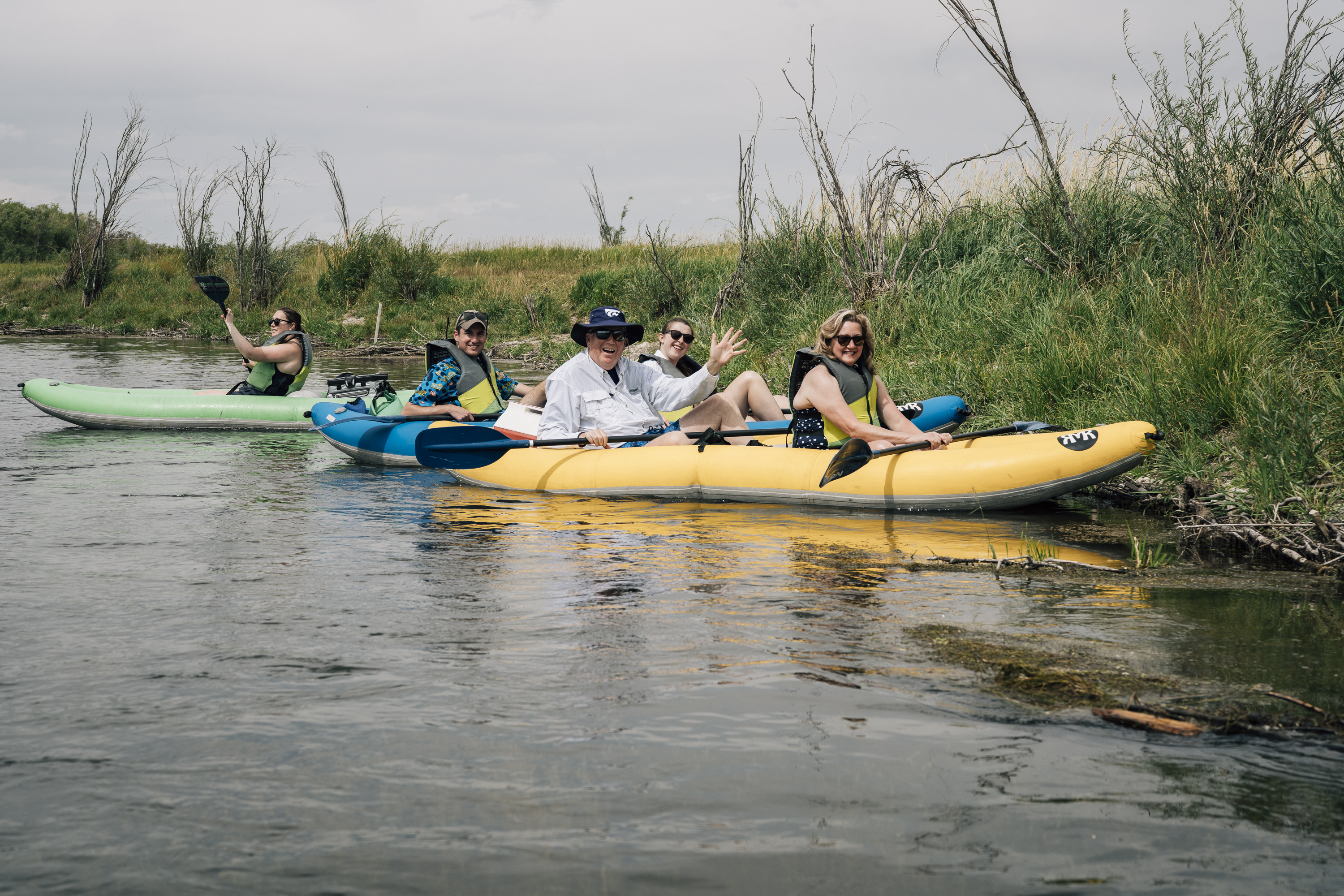 A group of six people paddles colorful kayaks along a calm river bordered by tall marsh grasses under an overcast sky