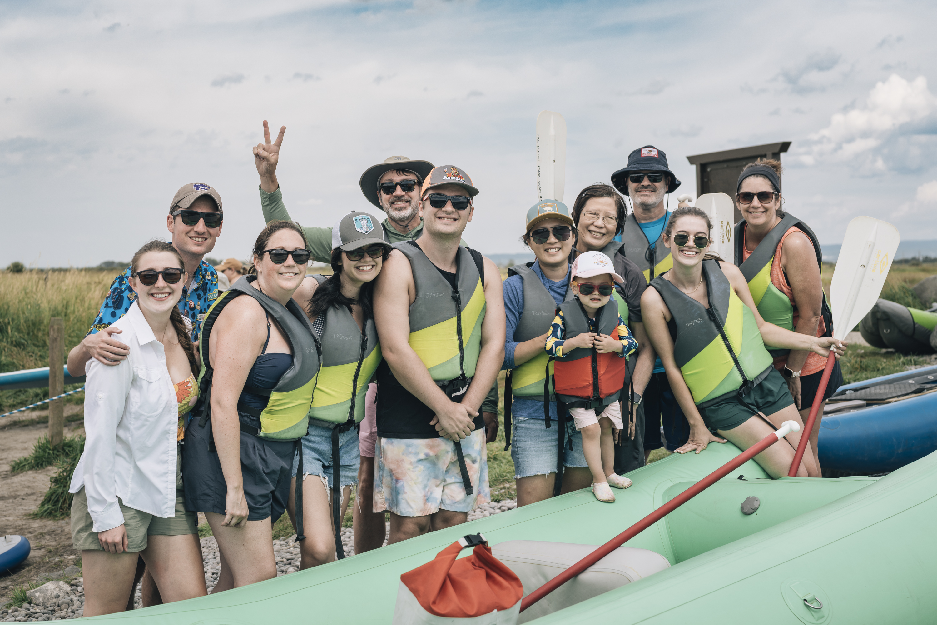 A group of twelve people wearing life vests and sunglasses stand together beside green rafts on a sandy riverbank under partly cloudy skies, posing cheerfully for a whitewater rafting trip