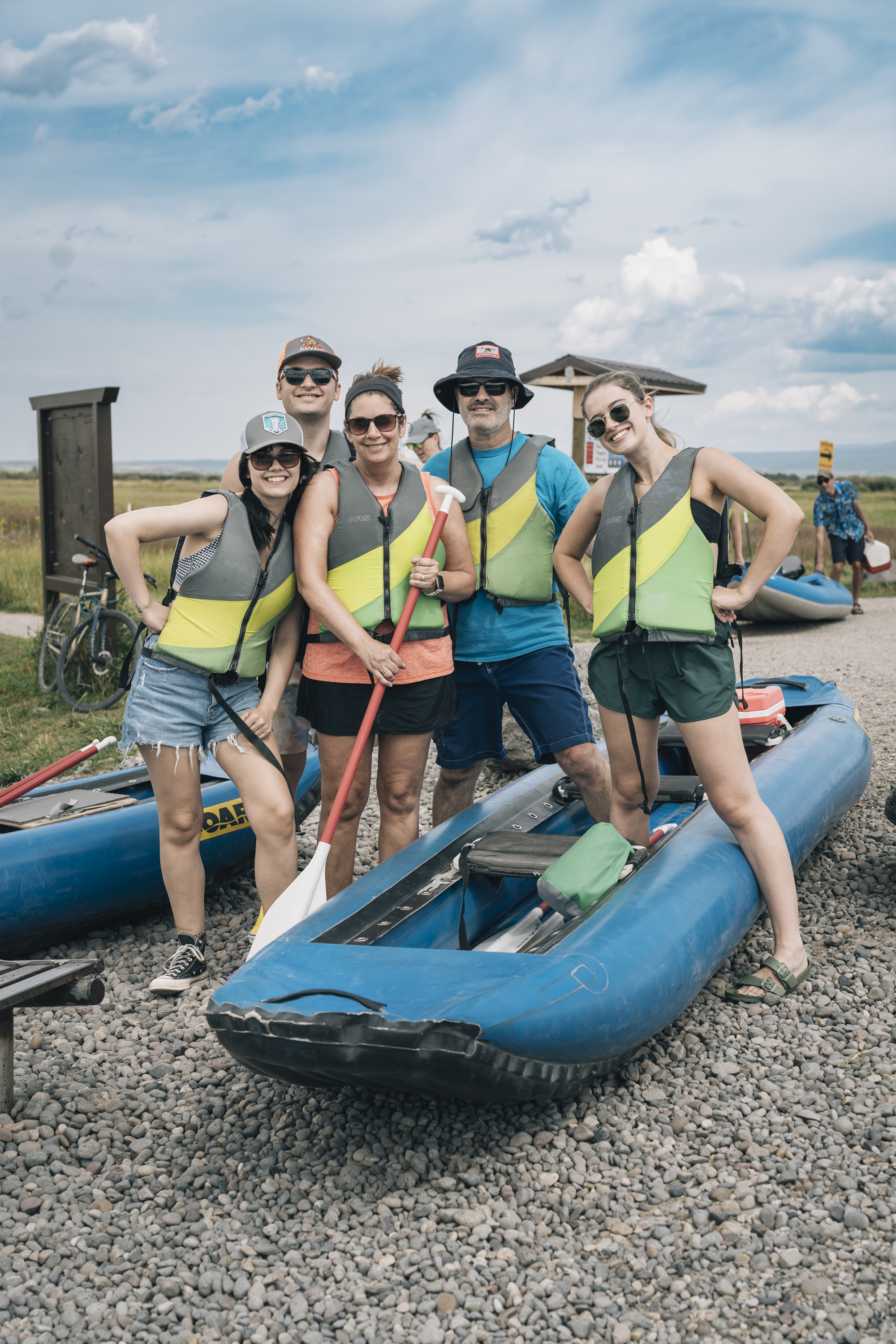 Five people wearing bright yellow and gray life vests pose beside blue inflatable rafts on a gravel riverside launch, with expansive plains and cloudy sky stretching behind them