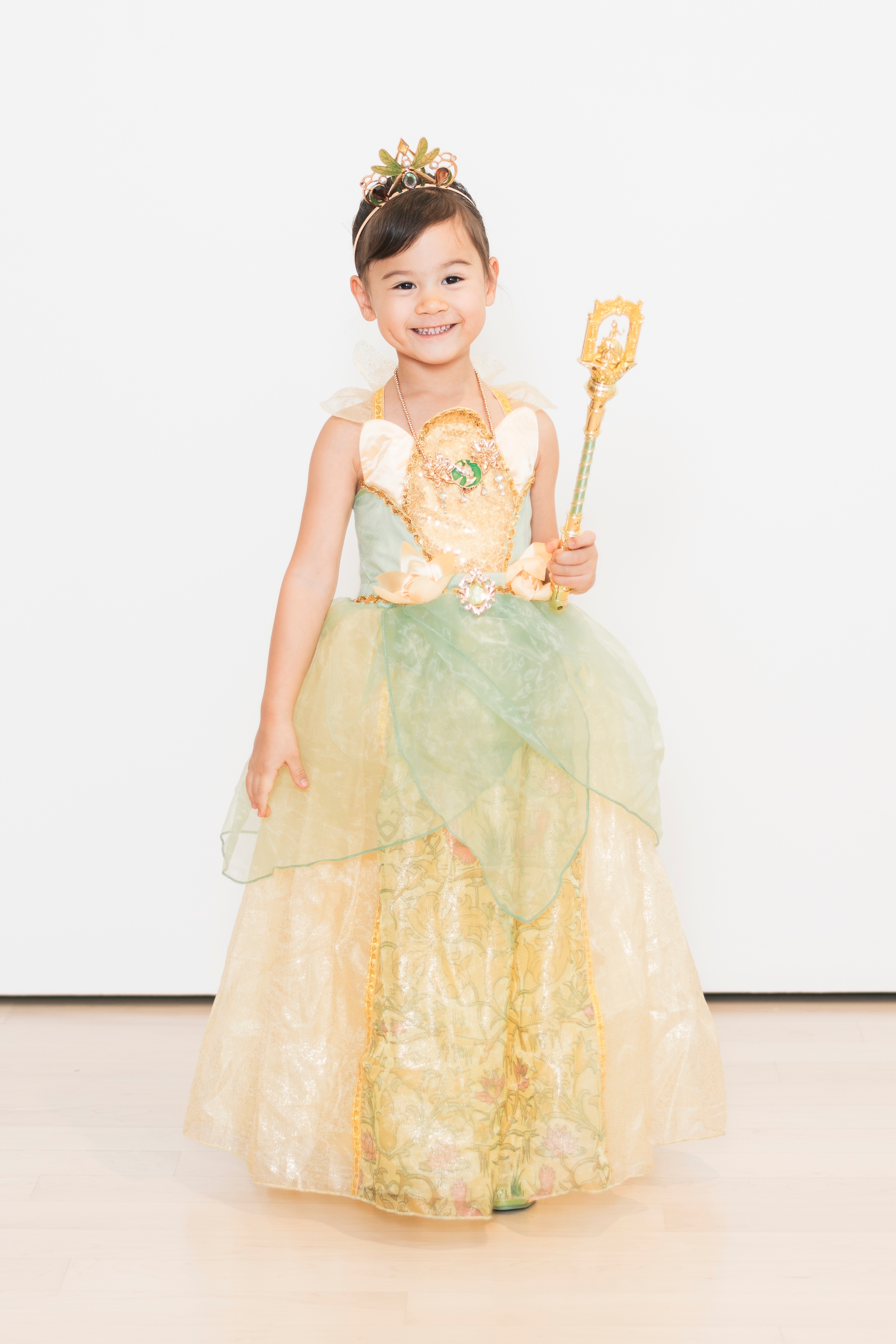 Young child dressed as Princess Tiana in gold and green gown, tiara, and holding ornate scepter against plain white background