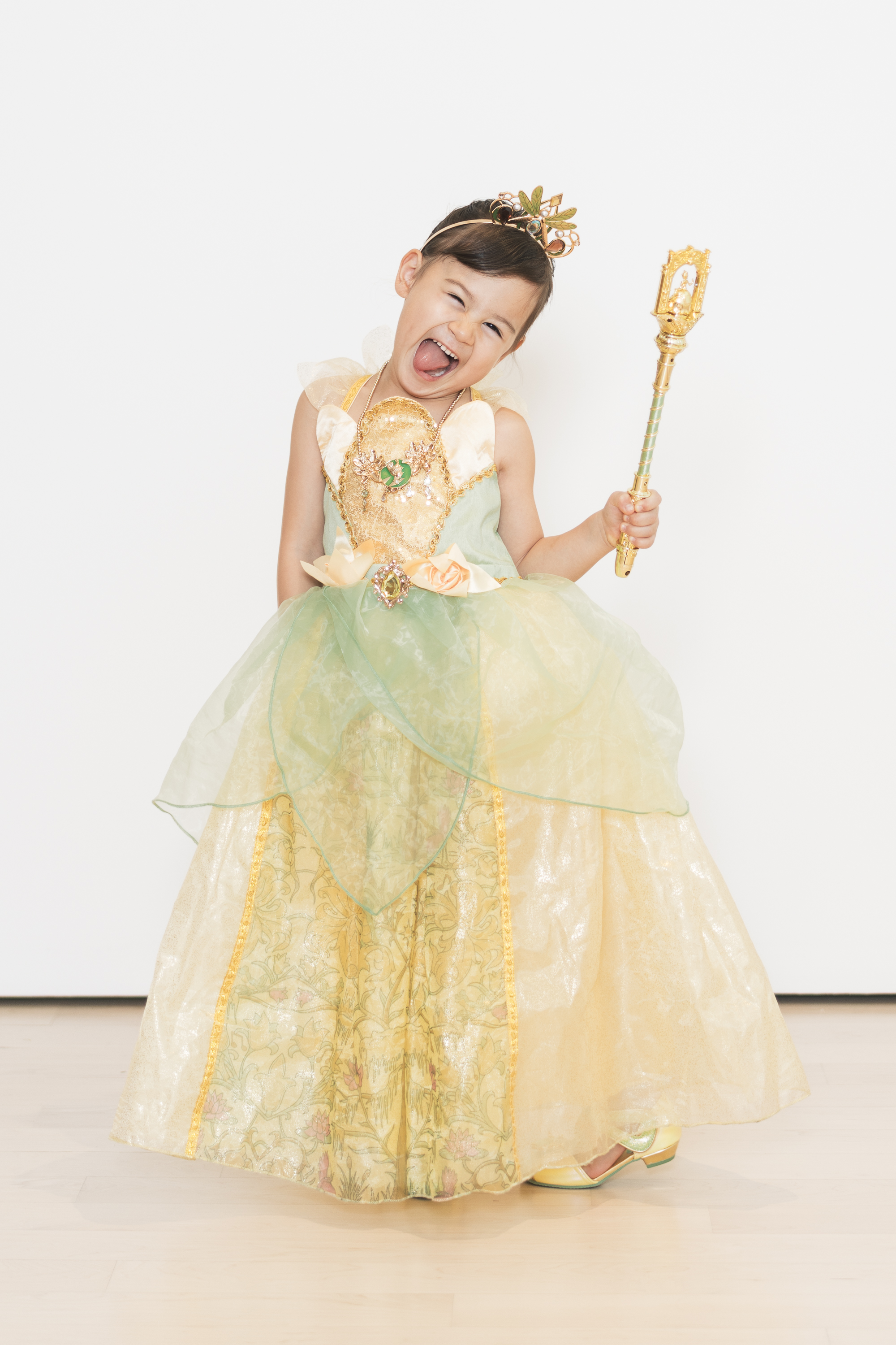 Young child dressed as Princess Tiana in an elaborate gold and green gown, tiara, and holding a jeweled scepter, poses playfully against a white backdrop