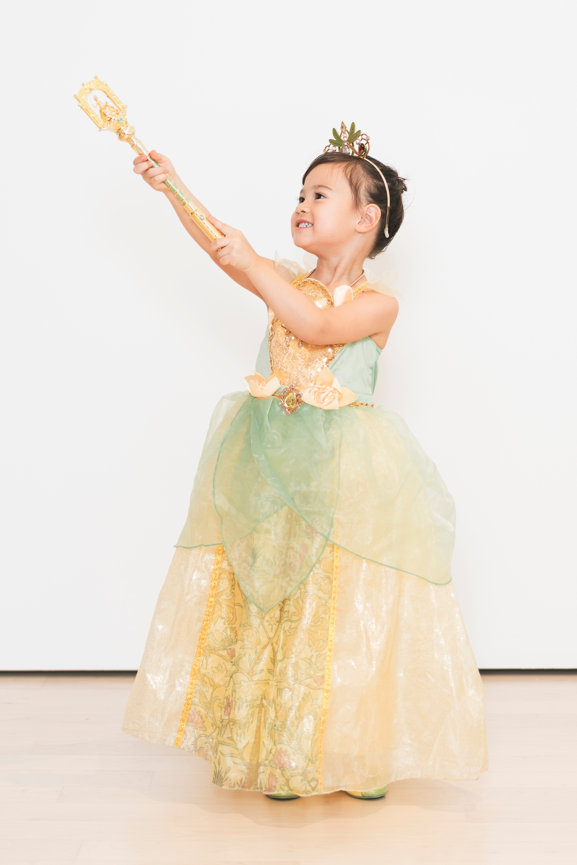 A young girl dressed as Princess Tiana smiles brightly while wearing a yellow and green ball gown with a gold tiara against a neutral white background