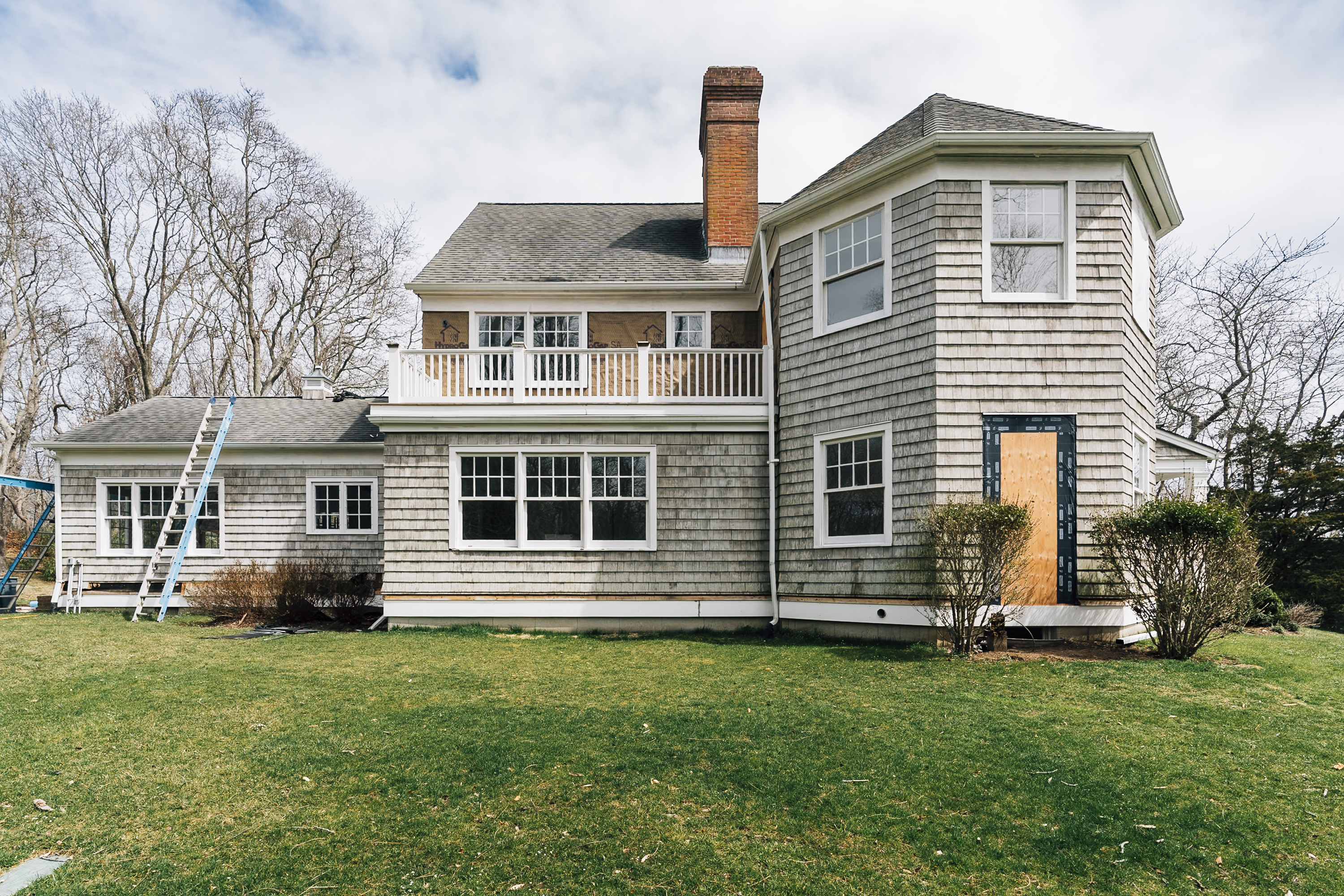 A two-story shingled house with white trim and a brick chimney sits on a manicured lawn, showing renovation work in progress with a ladder and boarded door visible