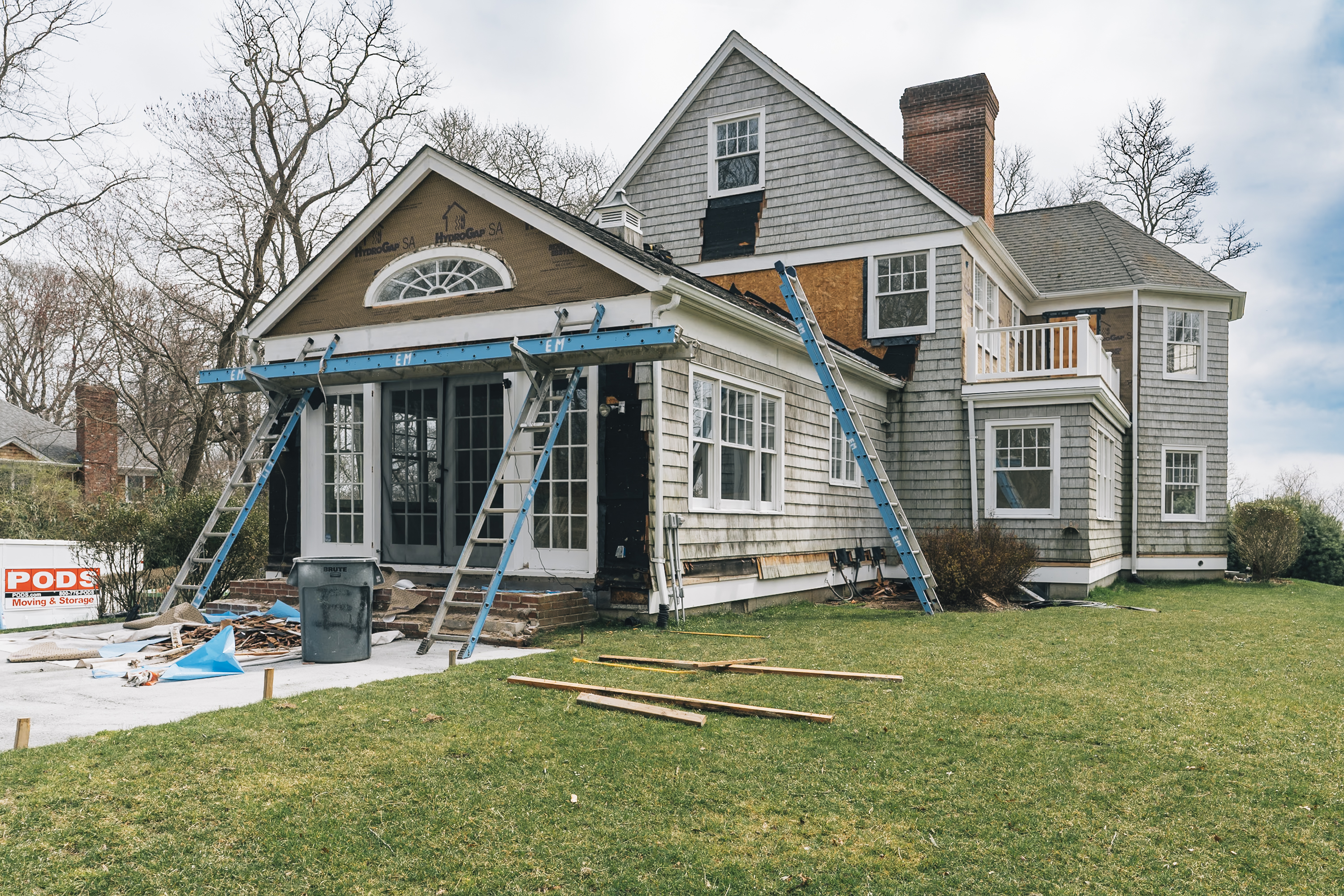 A traditional New England house under renovation displays blue scaffolding around its front addition, with bare trees and green lawn visible under overcast skies.