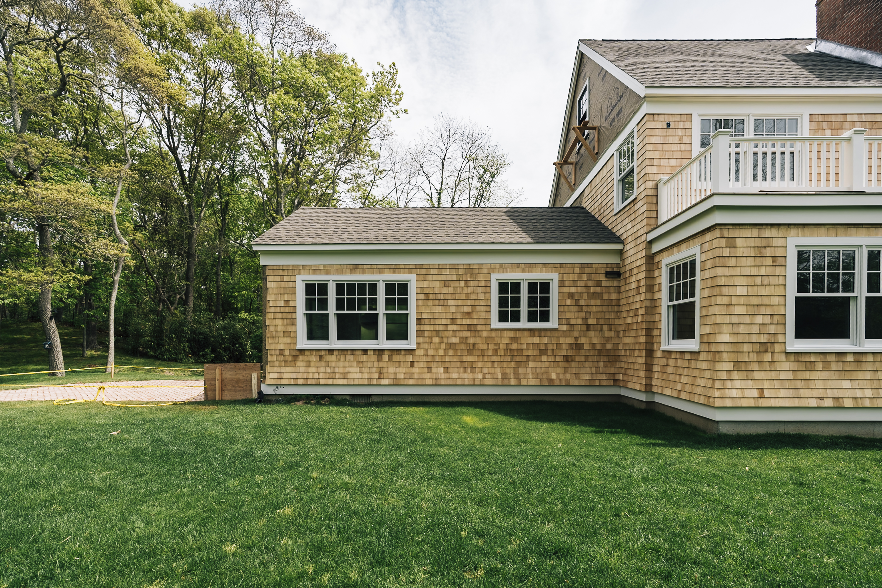 A cedar-shingled residential house with white-trimmed windows sits on manicured green lawn, backed by leafy trees under overcast spring daylight