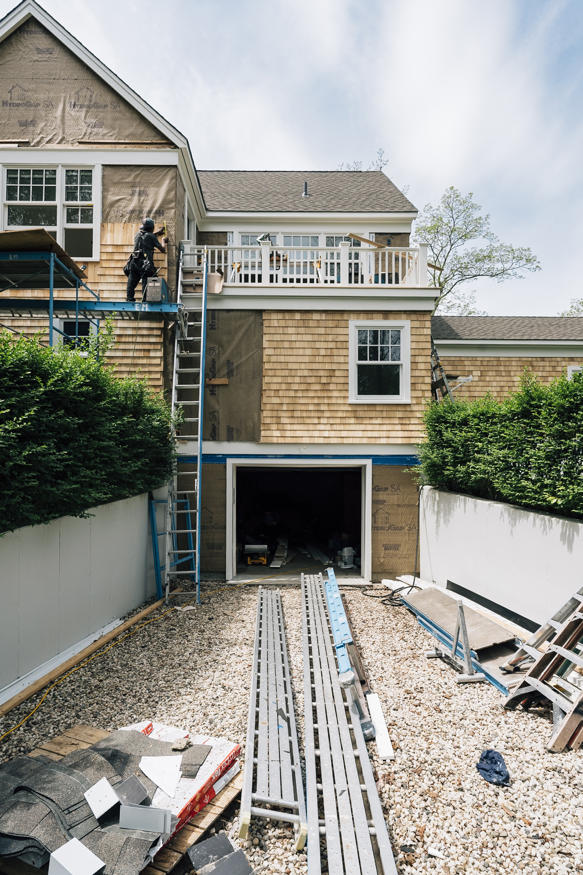 Shingle-clad residential house under renovation with scaffolding, open garage, gravel driveway, and construction materials visible in the enclosed courtyard space