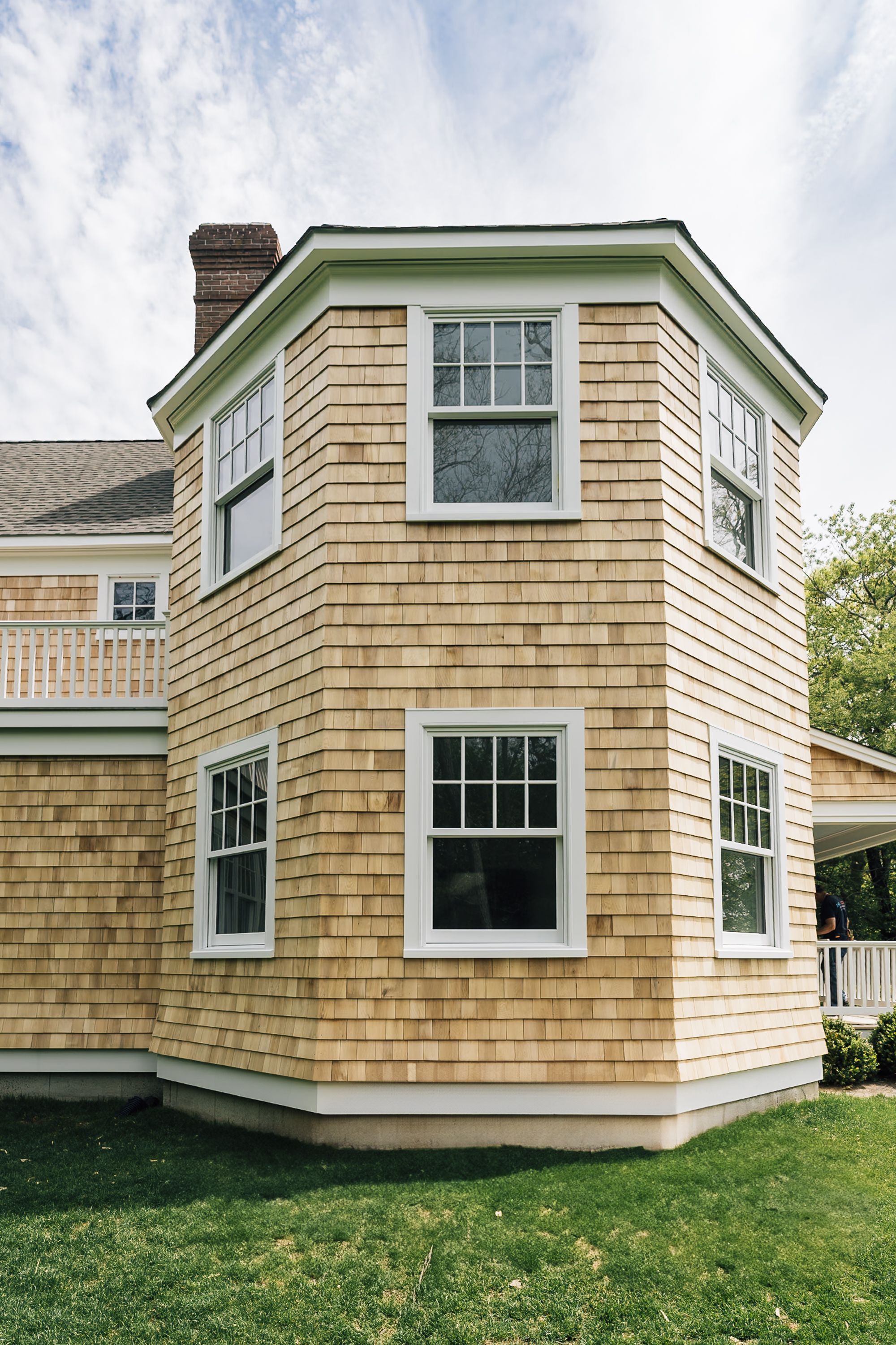 A tan cedar-shingled house with white trim and multiple windows stands against a cloudy sky, featuring a distinctive rounded tower section and manicured lawn in the foreground