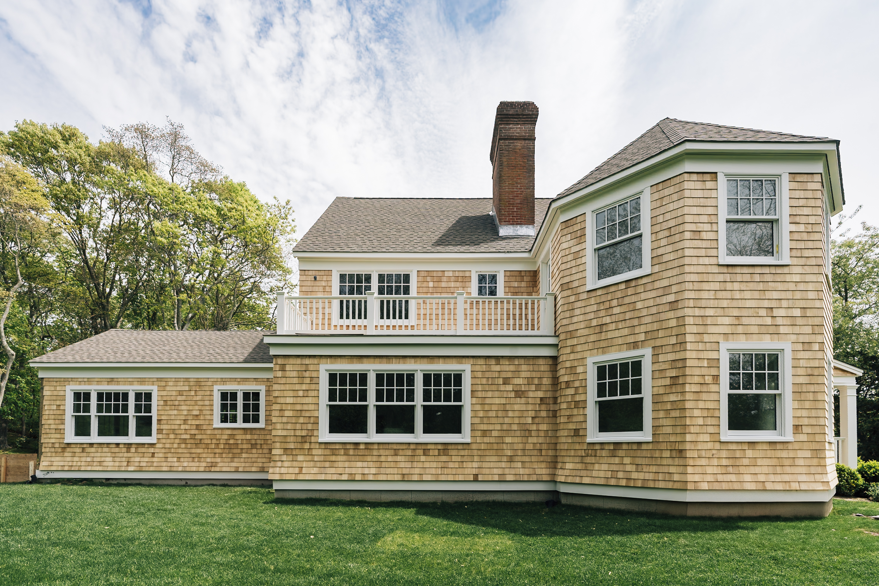 A multi-story residential home with tan cedar shingles and white-trimmed windows sits on a manicured lawn under partly cloudy skies, featuring a brick chimney and covered deck