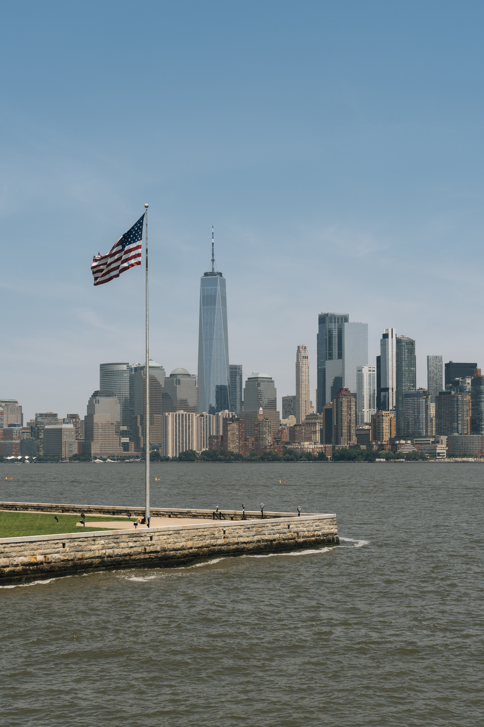 An American flag waves from a waterfront pier with the Manhattan skyline stretching across the horizon under clear blue sky and soft daylight