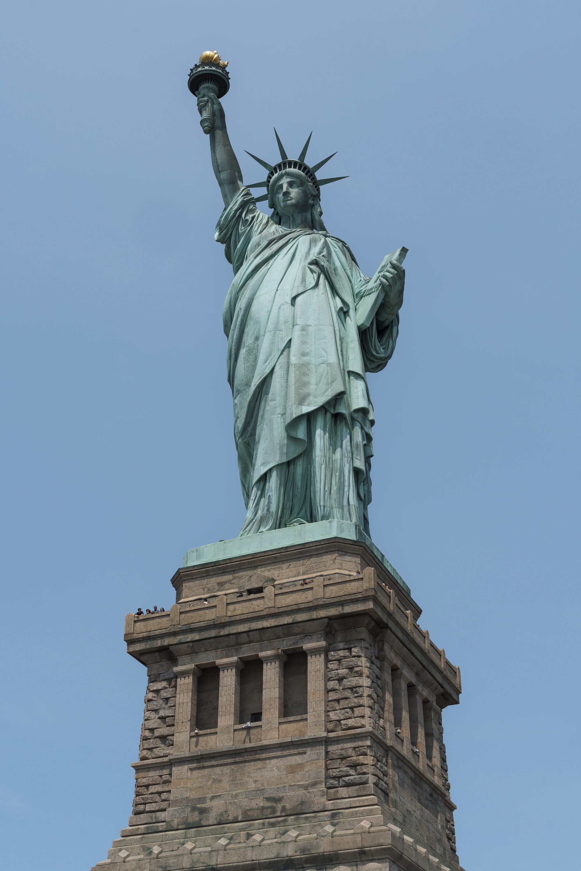 The Statue of Liberty stands against a clear blue sky, its copper patina visible on the upraised torch-bearing arm and spiked crown, mounted atop a massive stone pedestal with classical columned architecture