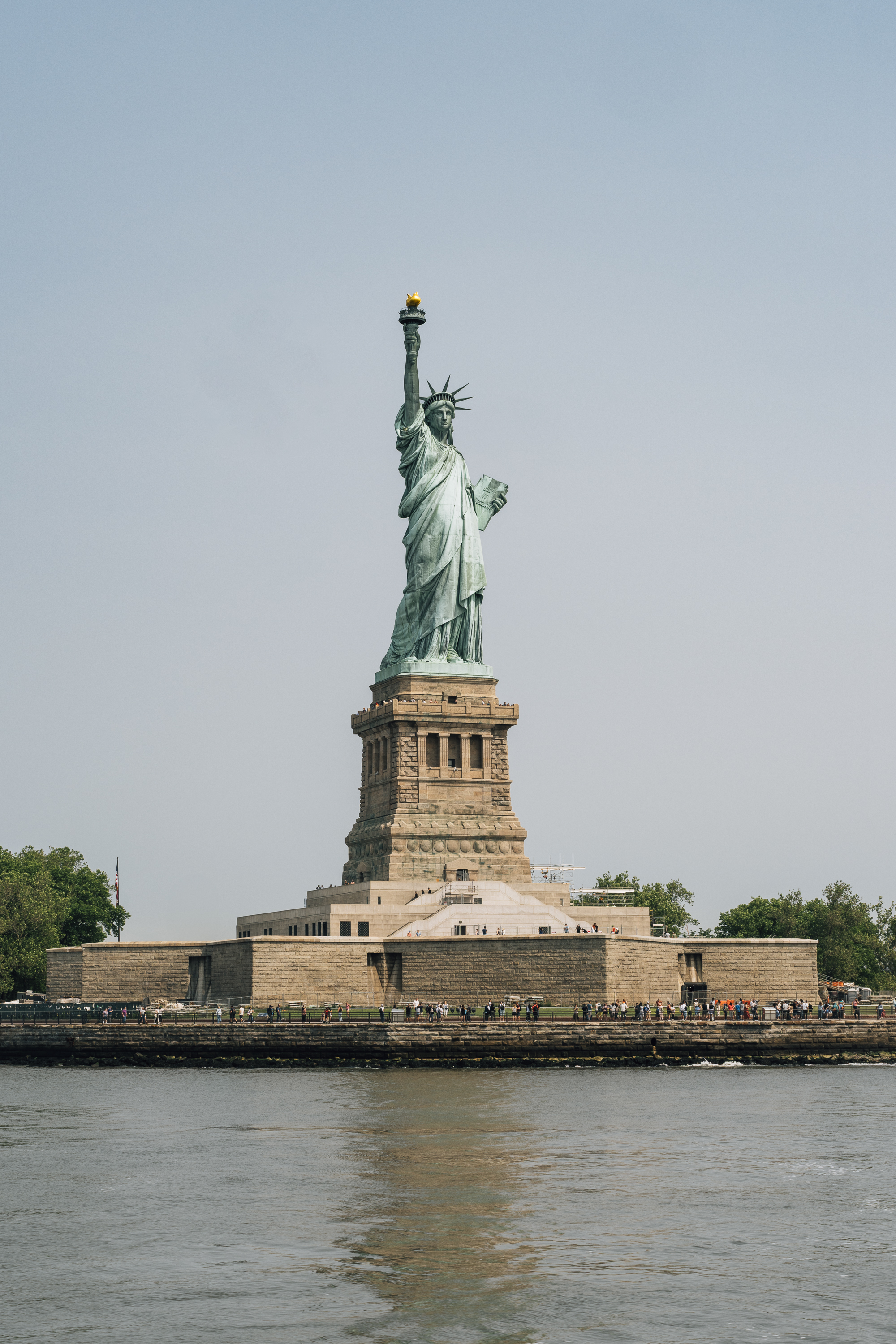 The Statue of Liberty stands on its stone pedestal on Liberty Island, viewed from the water with visitors gathered at its base under clear skies