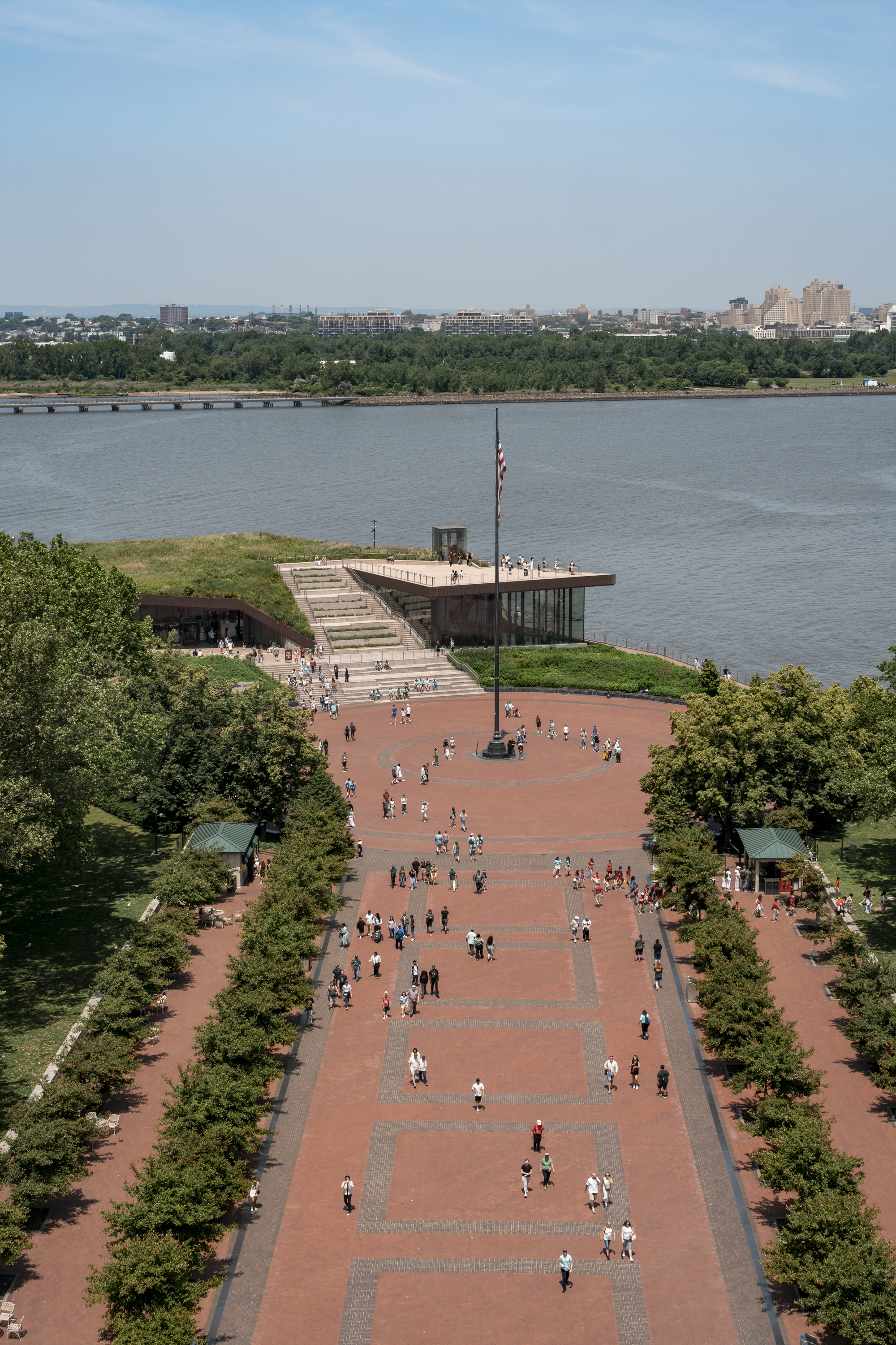 A brick promenade lined with green trees leads down to a modern waterfront pavilion overlooking the Hudson River, with Manhattan's skyline visible across the water under clear skies
