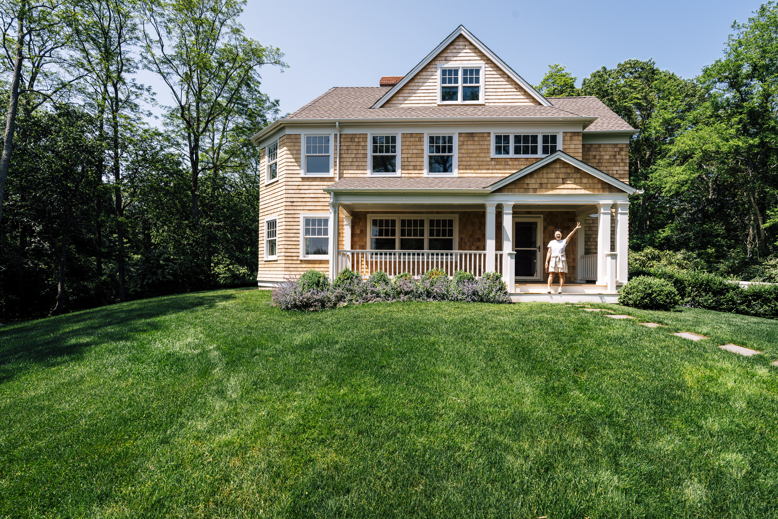 A two-story colonial-style house with mixed shingle and vinyl siding sits on a manicured lawn, framed by mature trees under clear blue sky