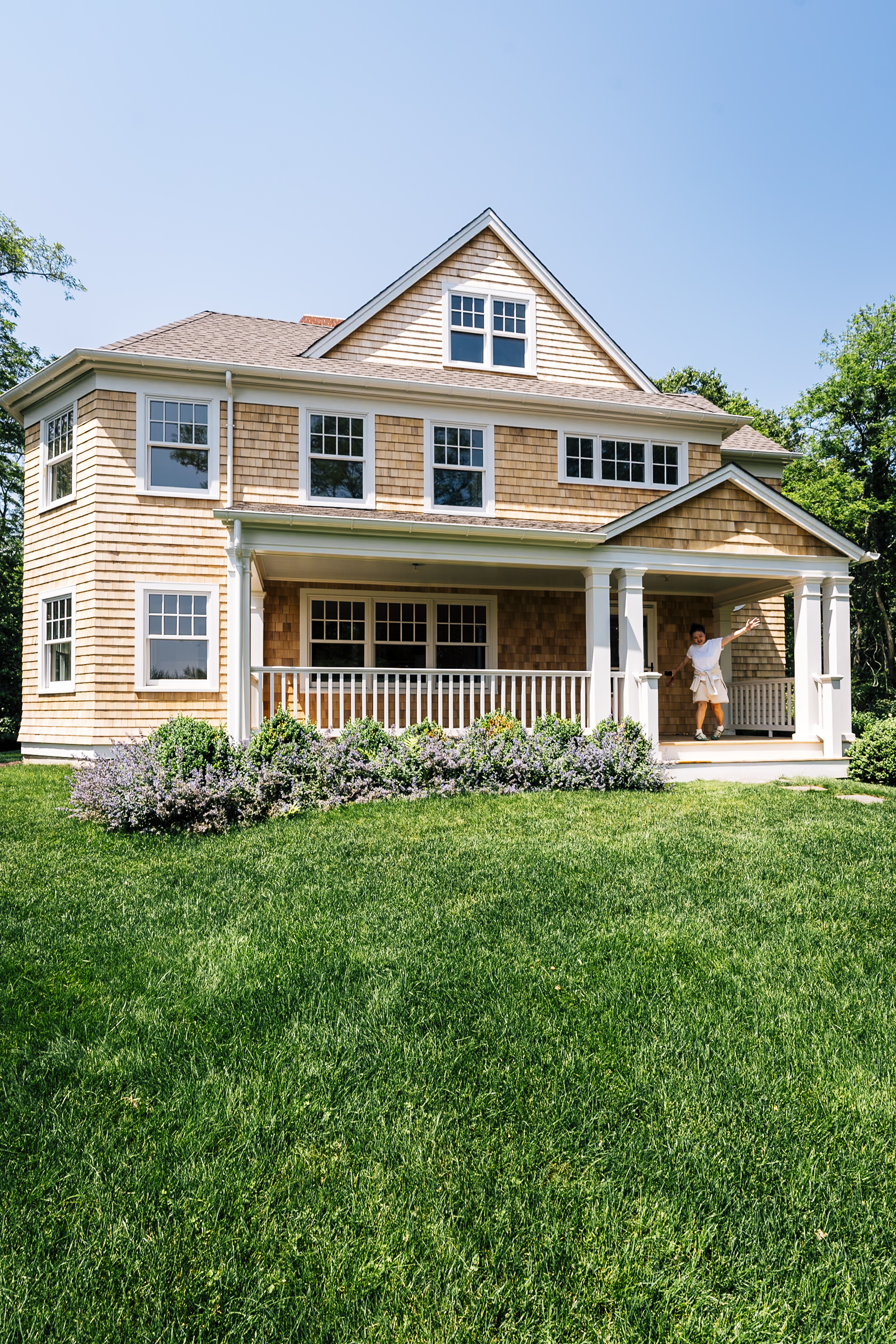 A two-story shingled colonial home with white trim and columns sits behind a manicured lawn, featuring a wraparound porch and gable roof under clear blue sky