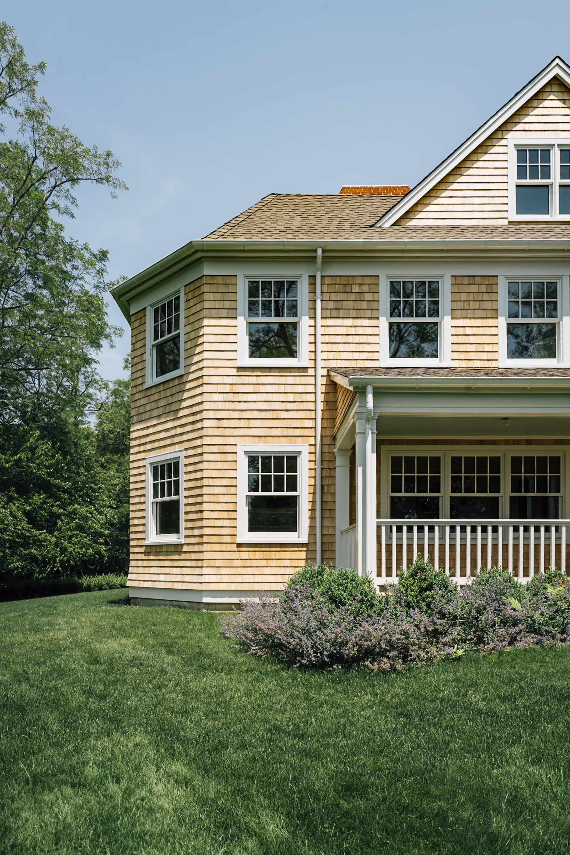A two-story colonial-style house with cedar shingle siding and black-trimmed windows stands on a manicured lawn under clear blue sky