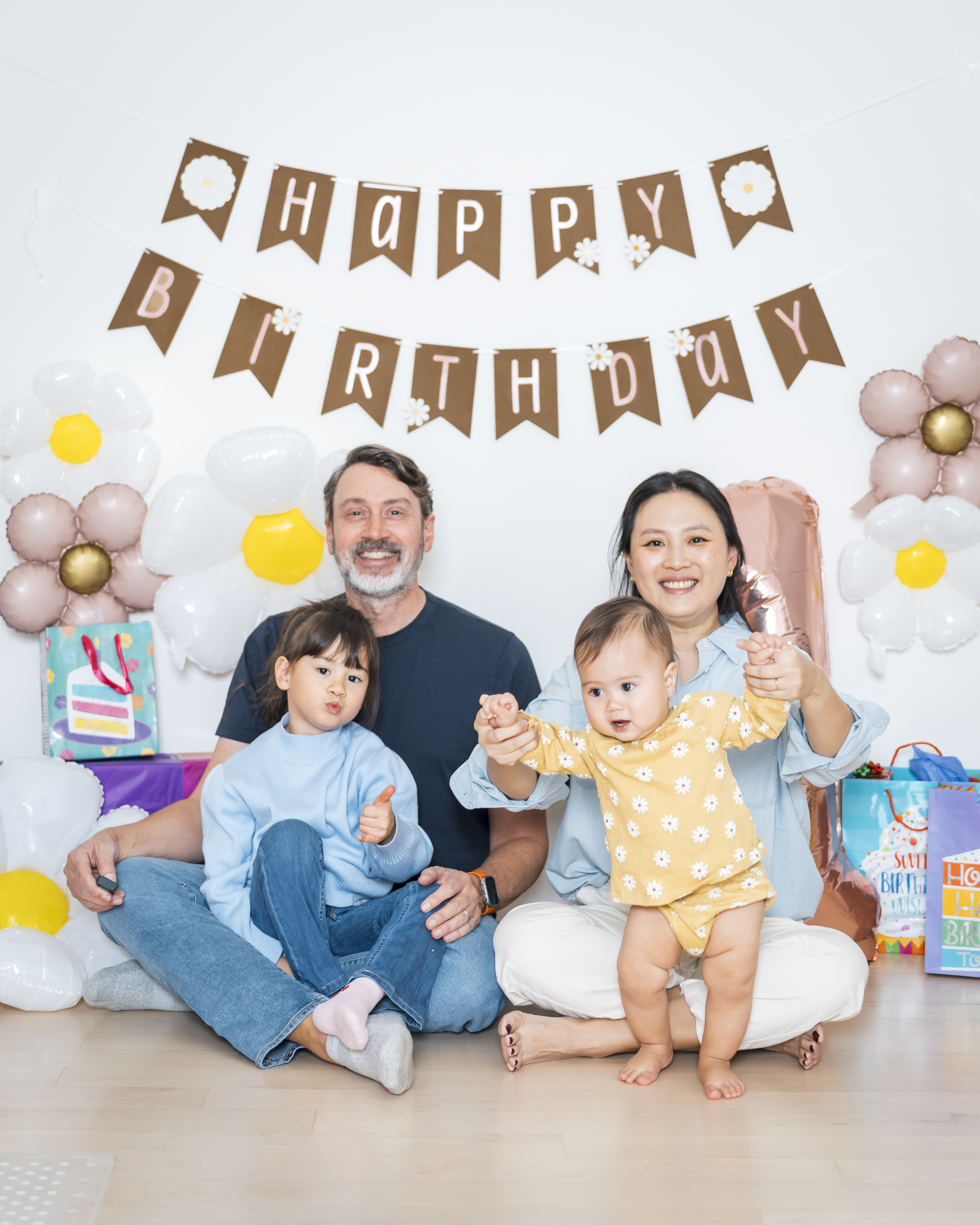 A family of four sits on a light wooden floor in front of a white wall decorated with a brown "Happy Birthday" banner and pastel balloon flowers, surrounded by wrapped gifts