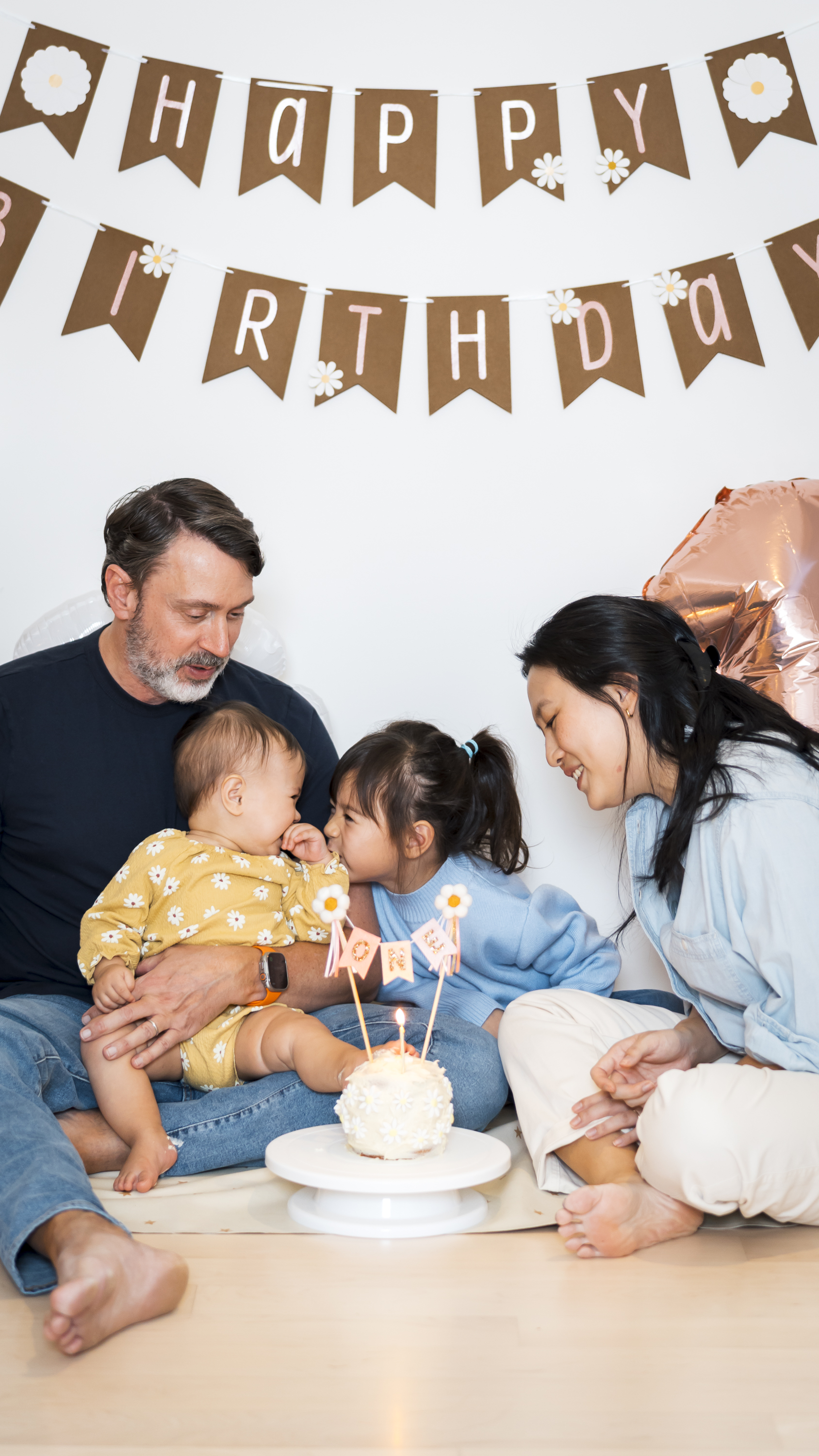 A family of four celebrates a child's first birthday around a small white cake, with brown and white bunting spelling "Happy Birthday" displayed above them