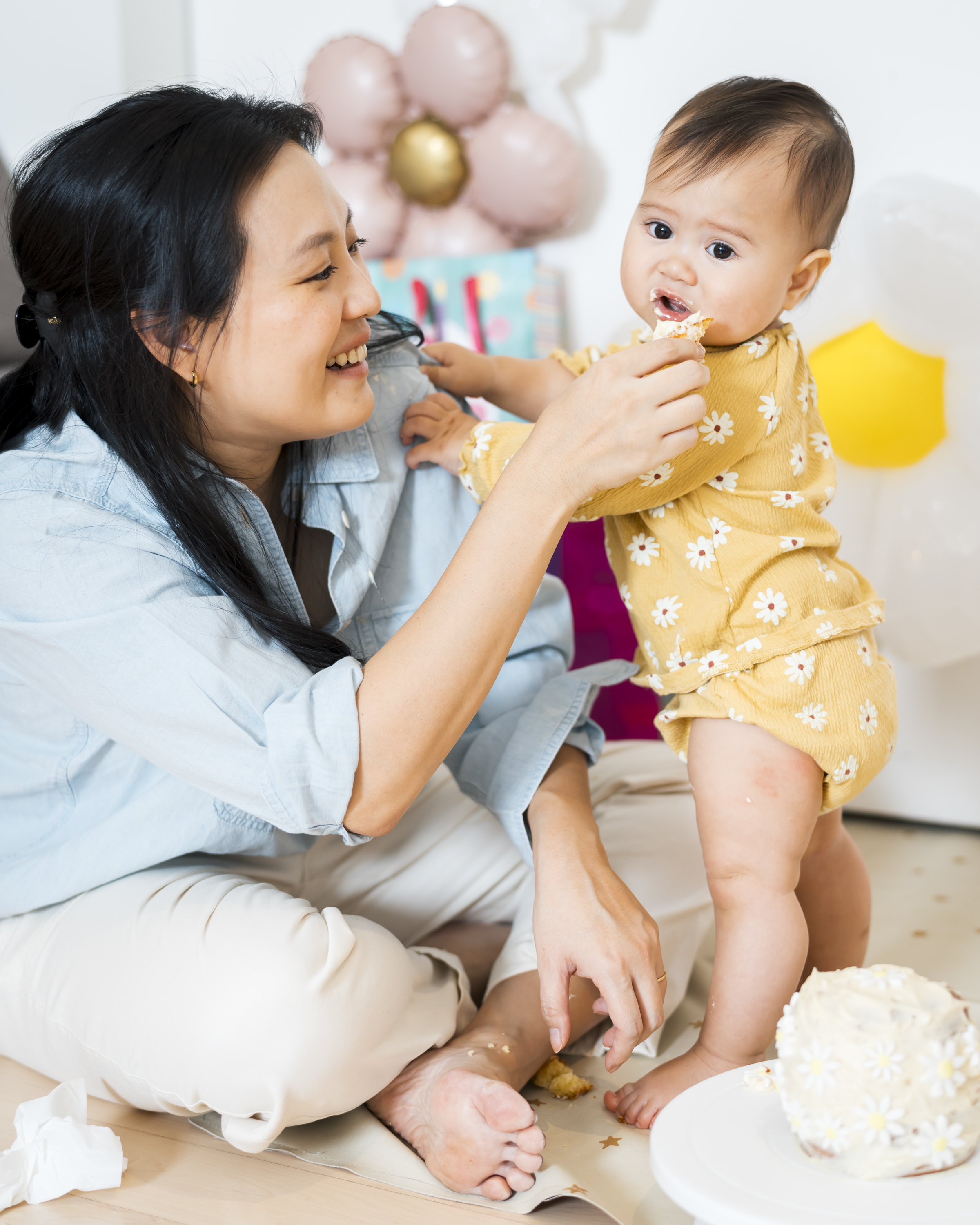 A woman in a light blue shirt playfully feeds cake to a smiling toddler in a yellow floral outfit during an indoor birthday celebration with pastel balloons in the background