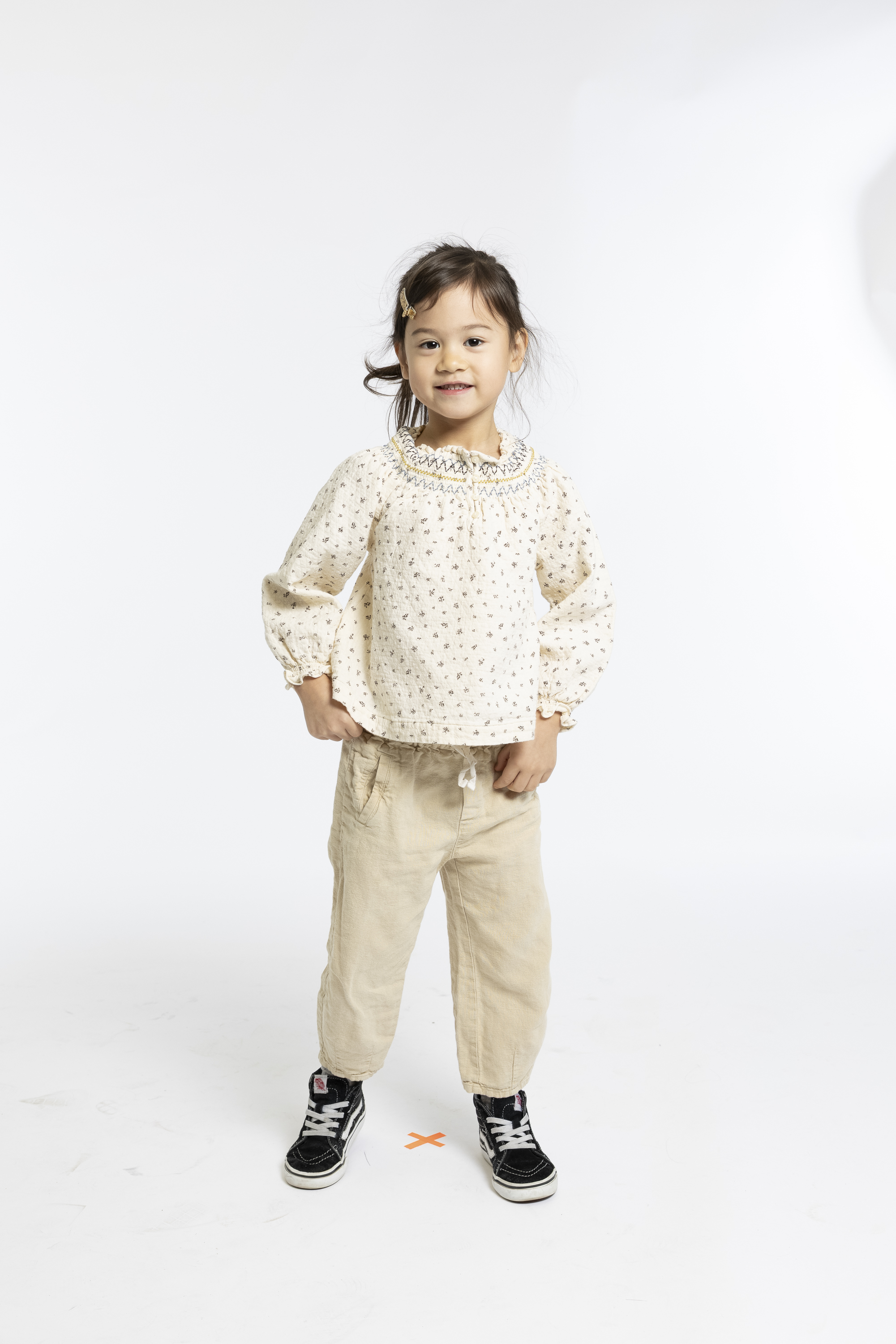 Young child stands confidently against a white backdrop wearing a cream polka-dot blouse, beige pants, and black-and-white sneakers with hands on hips