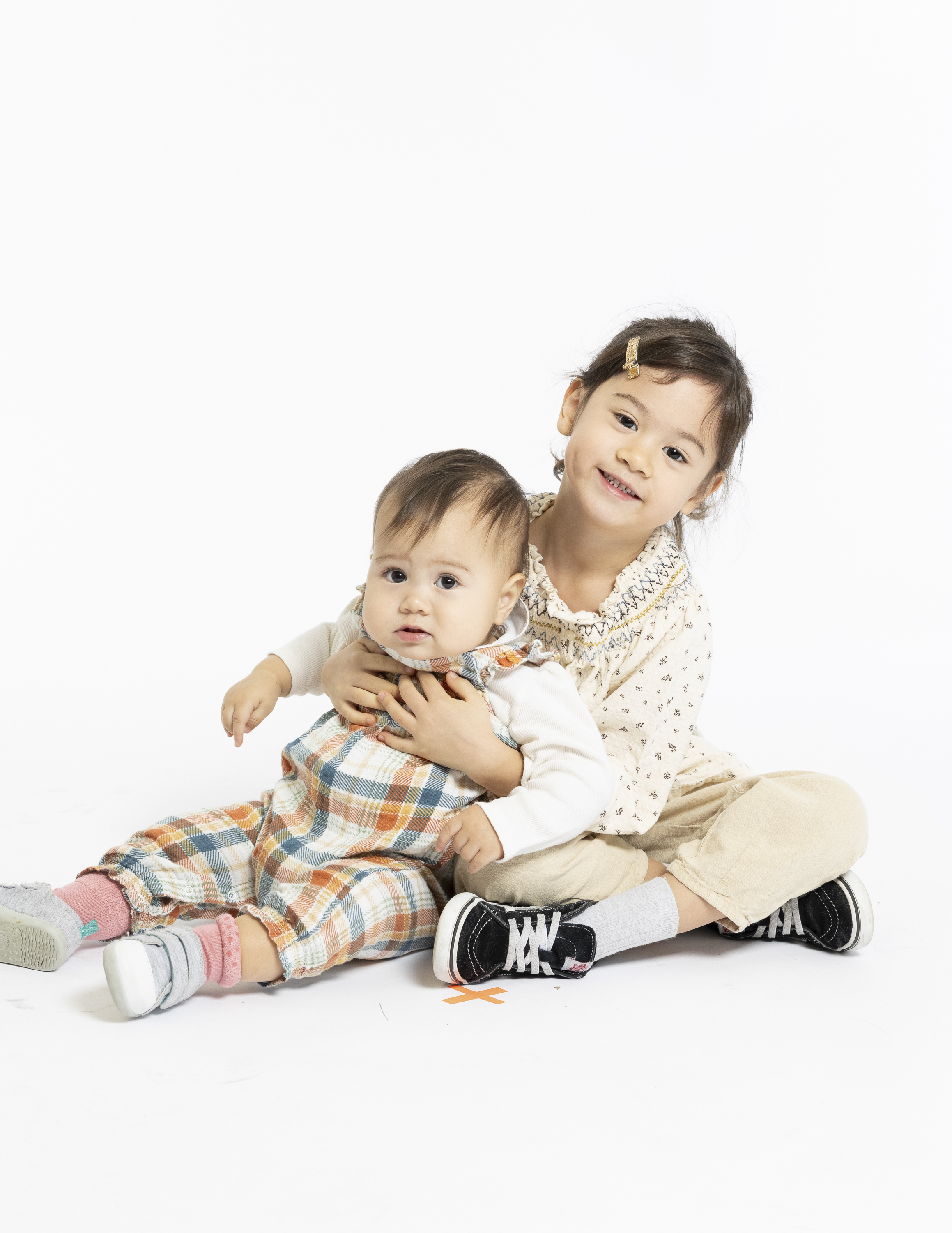 Two young children sit together on a white background, with an older girl in a cream patterned shirt hugging a younger toddler wearing plaid overalls and pink socks