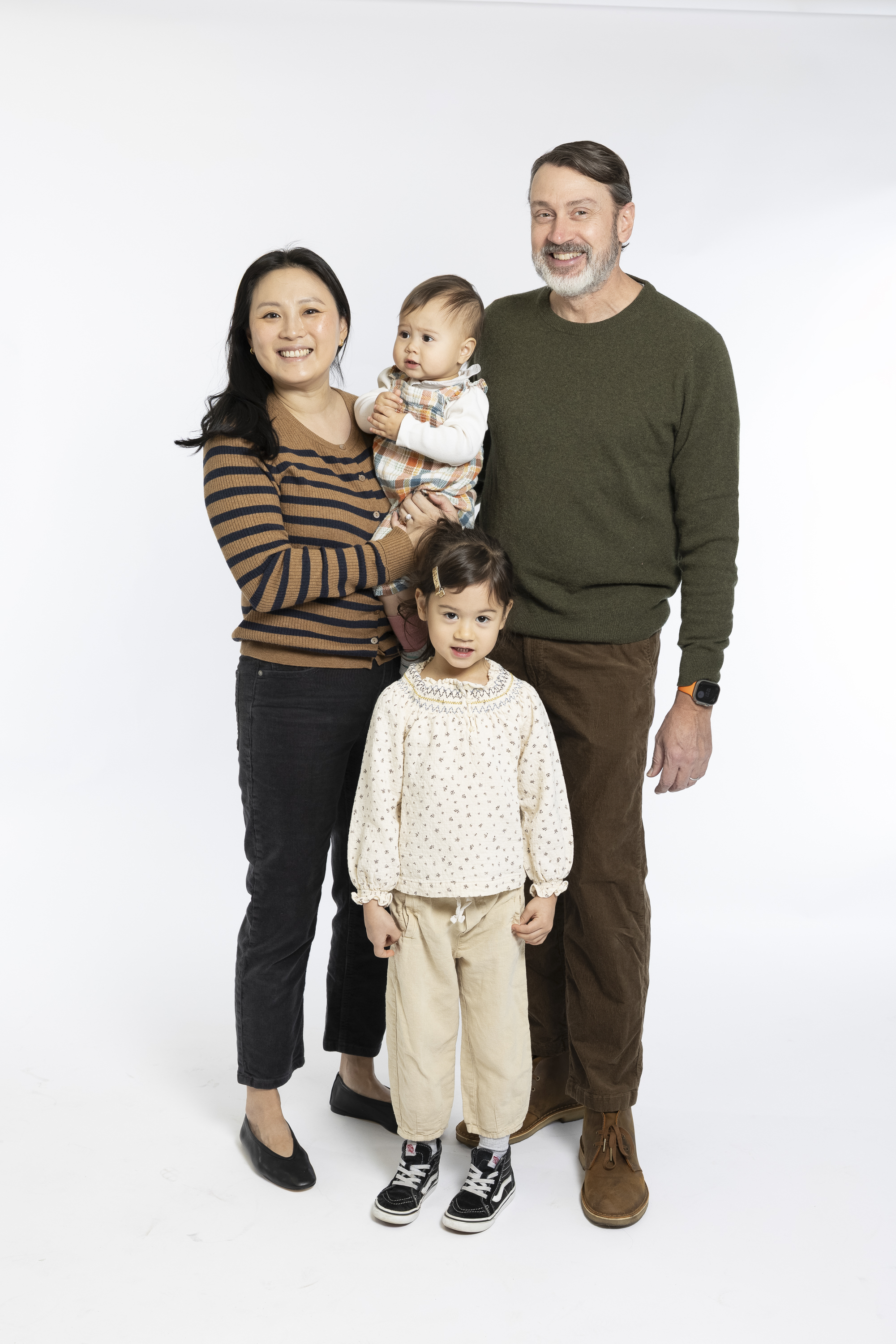 A family of four stands together against a white background, smiling at the camera with warm, natural expressions and casual clothing in neutral earth tones