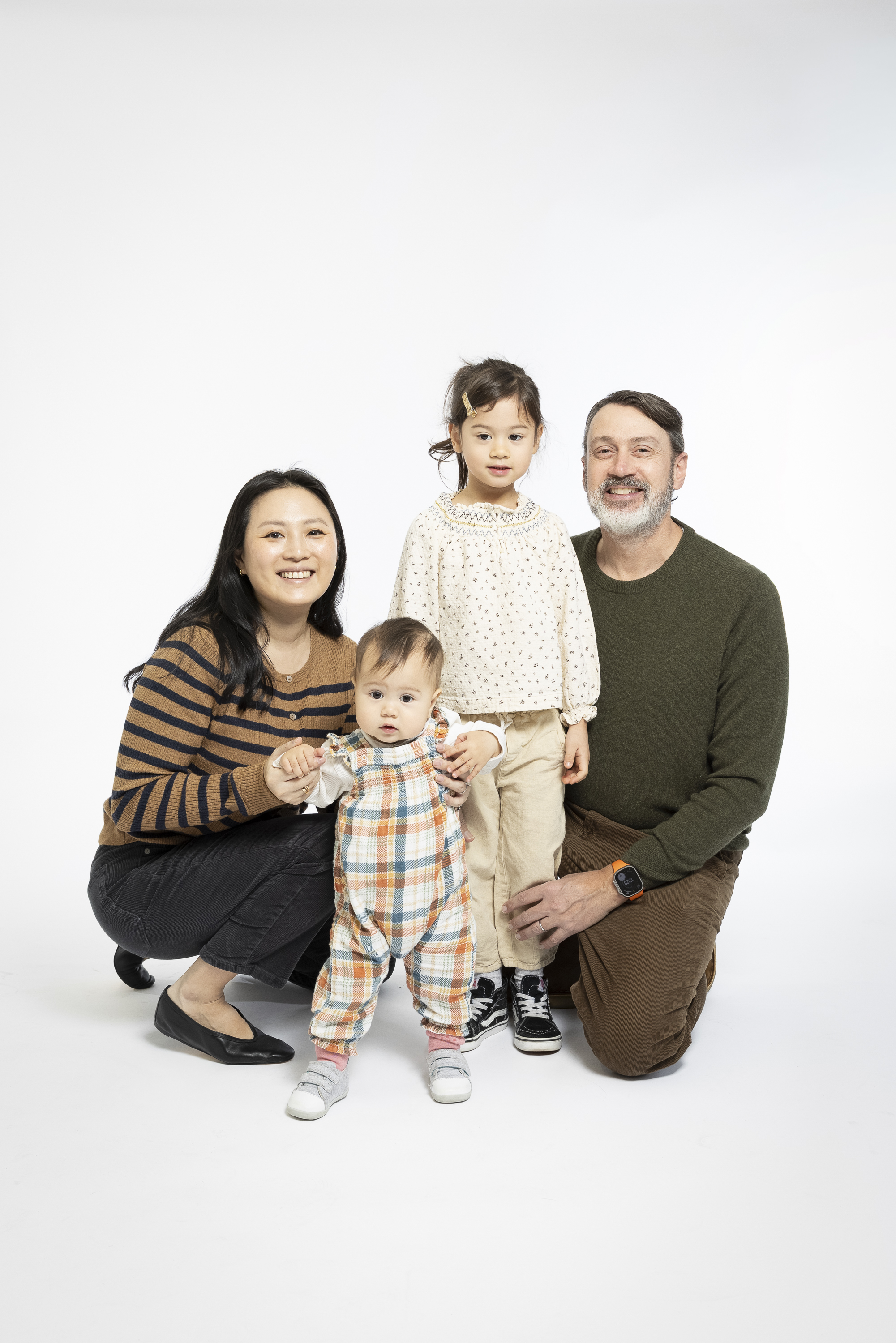 A family of four poses together against a plain white background, with two adults crouching beside a young toddler and an older child standing between them, all smiling warmly at the camera