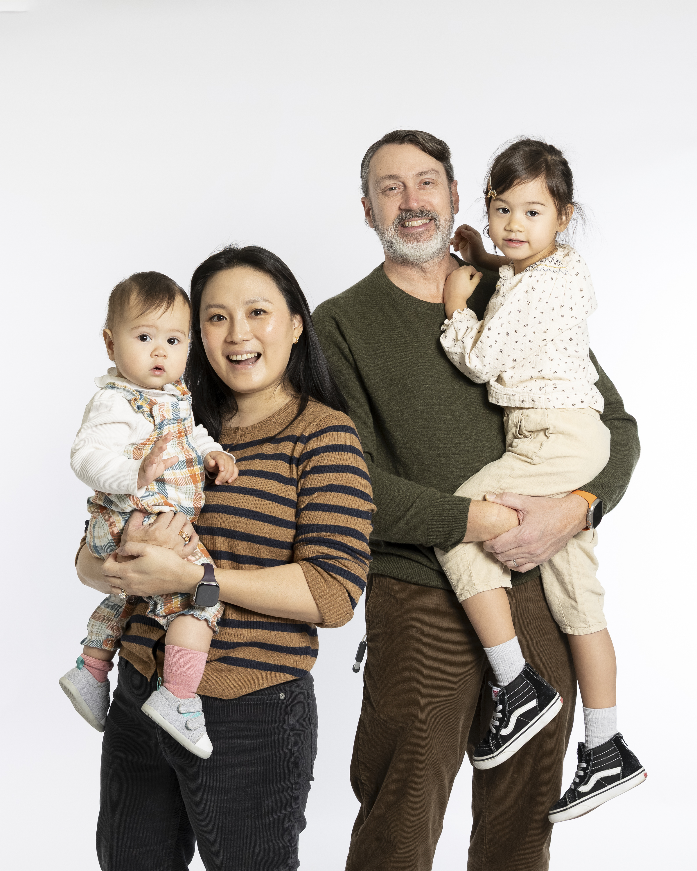 A family of four stands together against a white background, with two young children held in their parents' arms, all smiling warmly at the camera