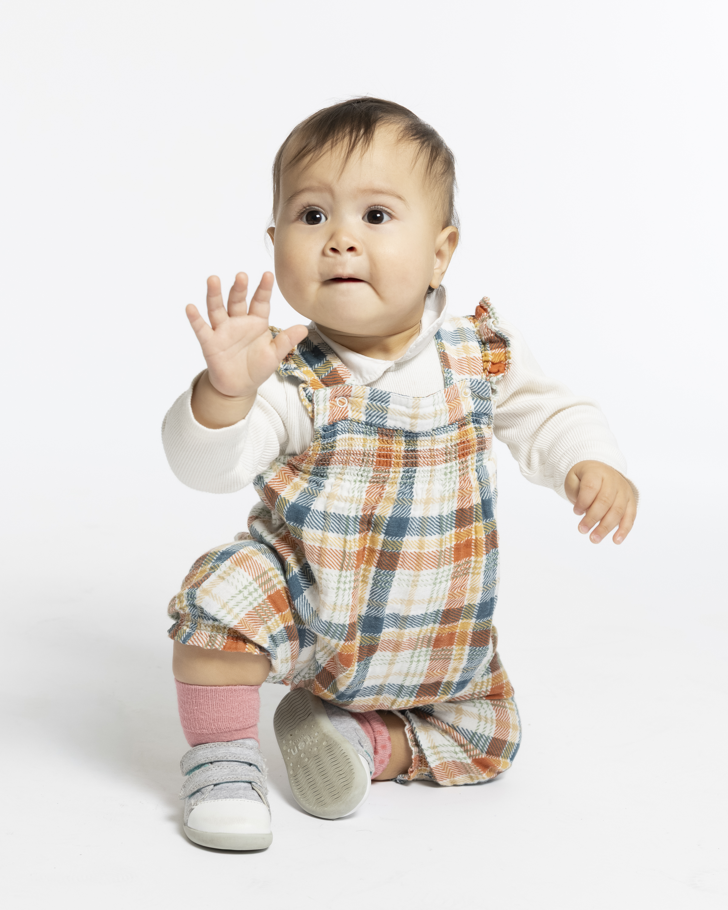 A toddler in plaid overalls and white long-sleeve shirt sits on a neutral background with one hand raised, wearing pink socks and white shoes