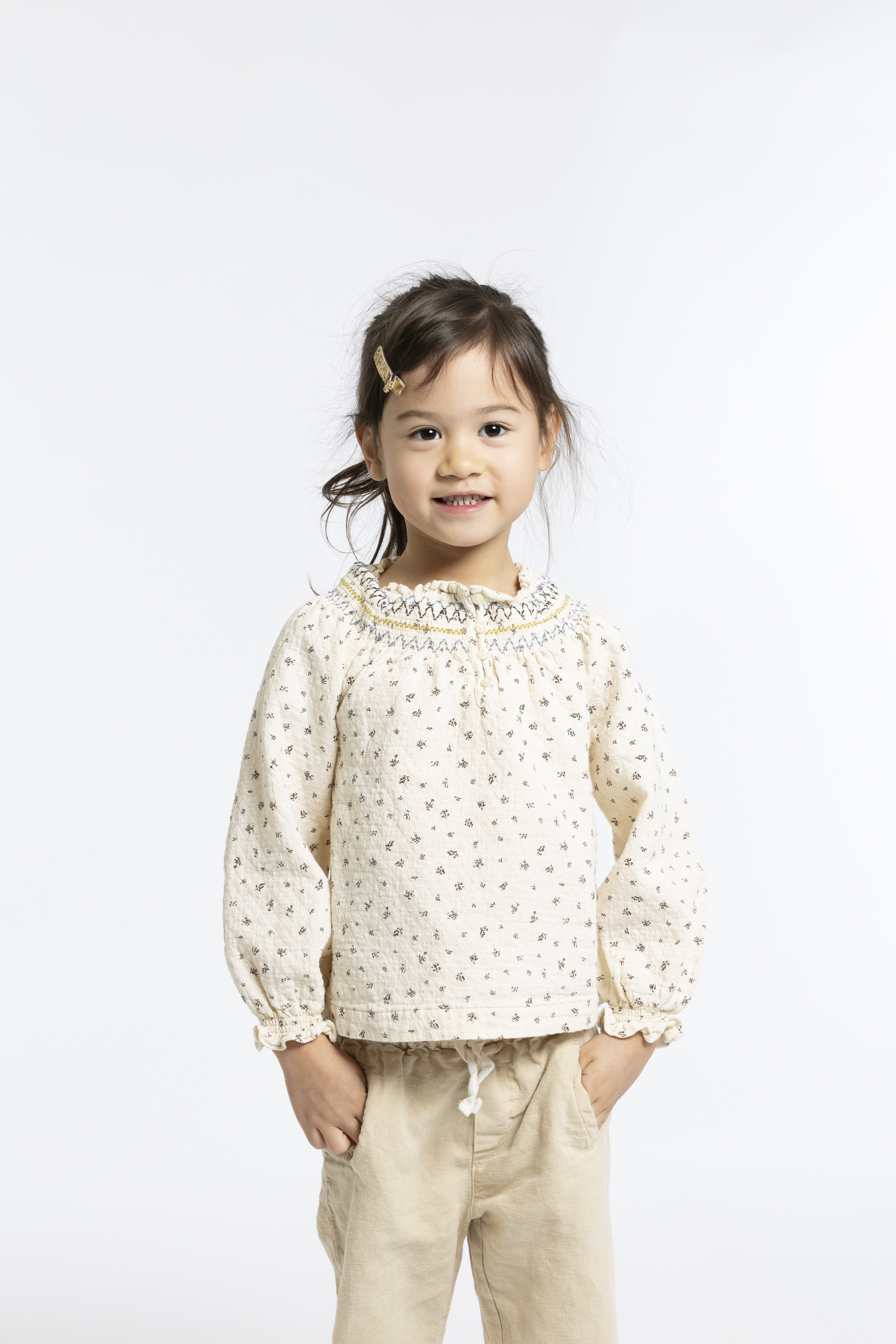 Young child with dark hair and a gold hair clip stands against a plain white background wearing a cream-colored patterned blouse and beige pants, smiling softly at the camera