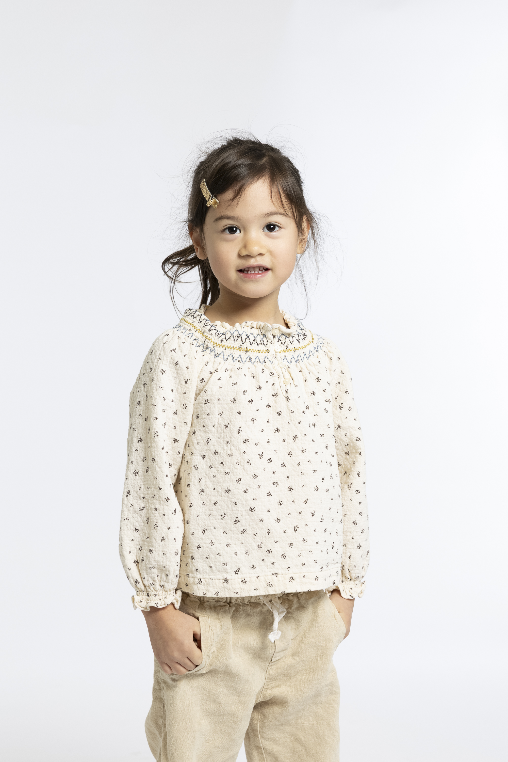 Young girl with dark hair and a gold hair clip wears a cream-colored floral blouse and beige pants against a plain white background, smiling softly at the camera
