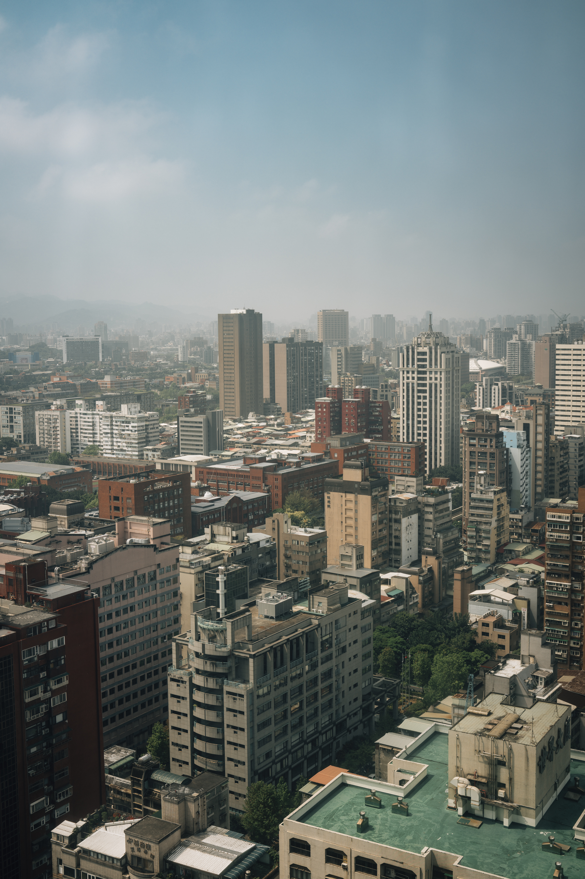 Dense urban skyline of Taipei stretches toward a hazy horizon under pale blue sky, with mixed-height buildings in brick, concrete, and glass dominating the composition