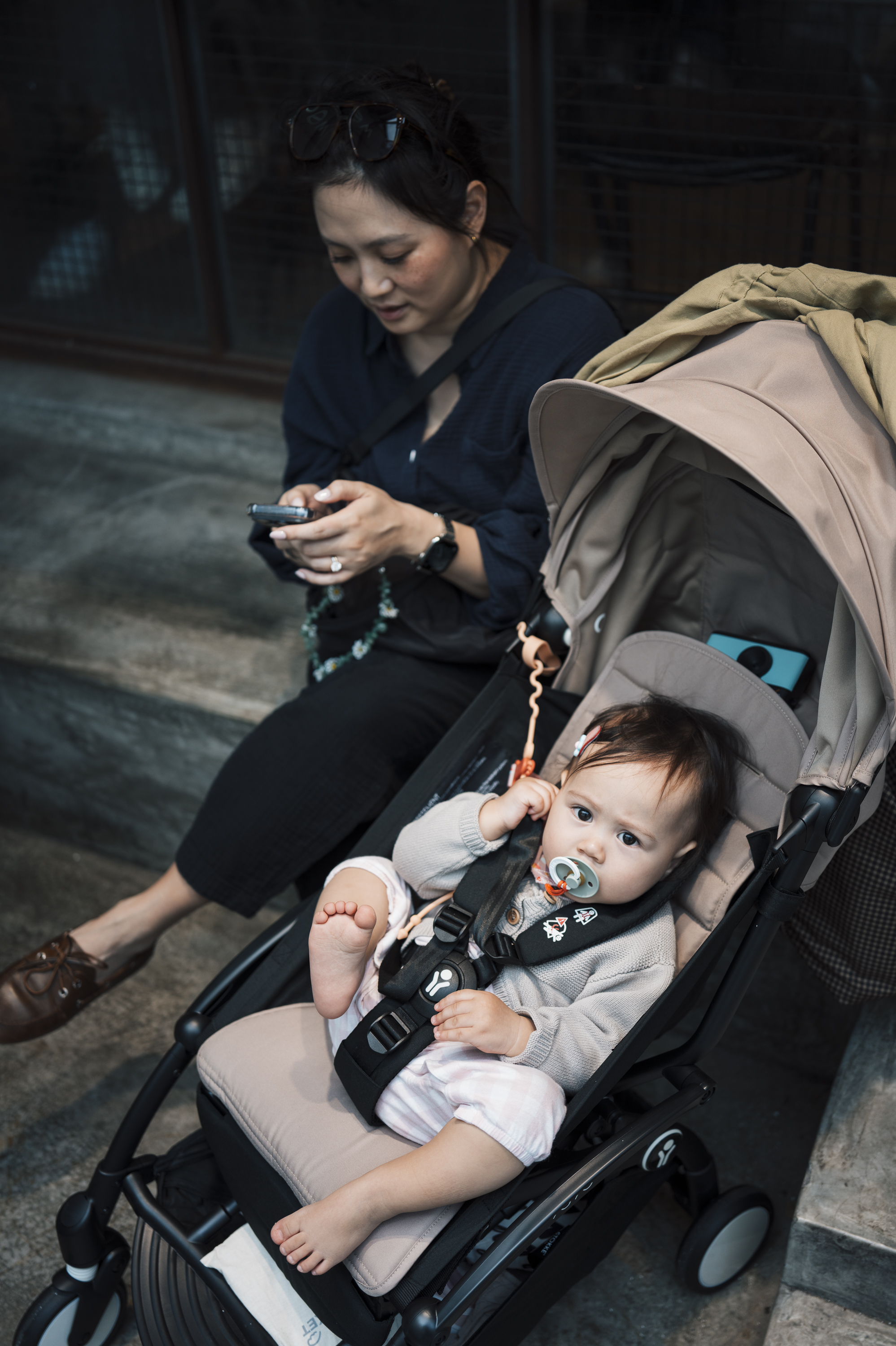 A woman in a dark navy shirt sits beside a gray stroller holding her phone, while a toddler in light clothing sits in the stroller on a concrete urban ground